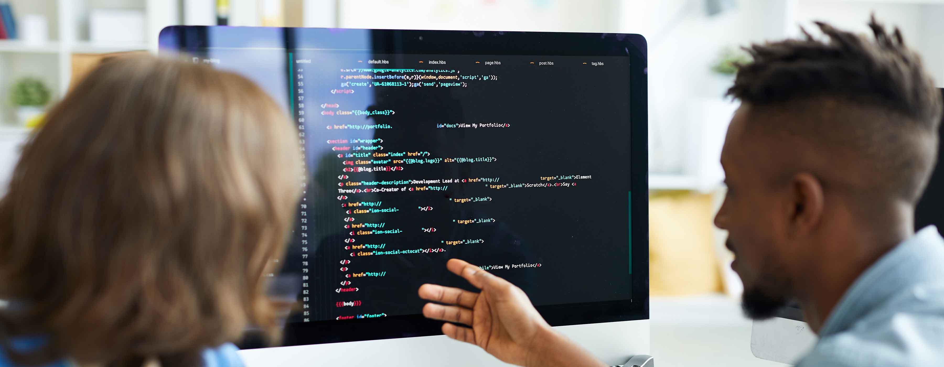 Foto de duas pessoas sentadas em frente a um monitor, analisando códigos de programação exibidos na tela.