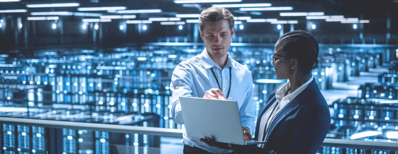 Foto de um homem mostrando dados em notebook para uma mulher em sala de servidores de empresa.