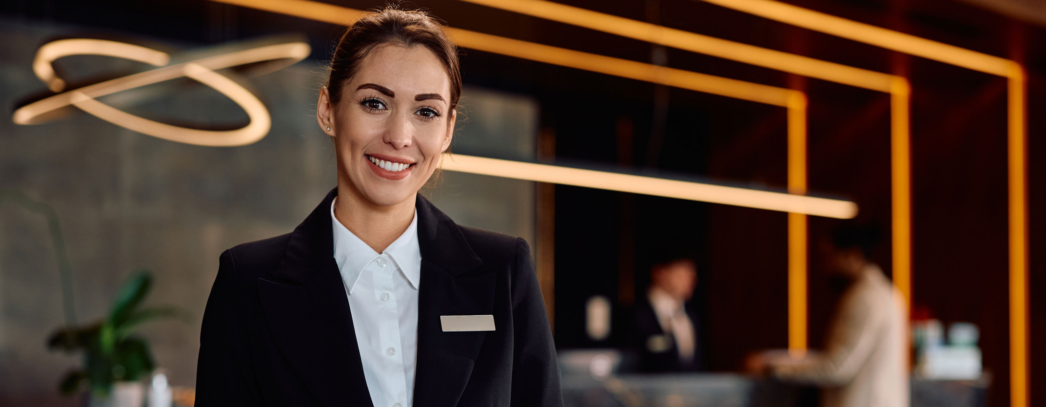 Foto de atendente de hotel sorrindo, usando uniforme social, em frente ao balcão de recepção, com fundo moderno e iluminação decorativa.