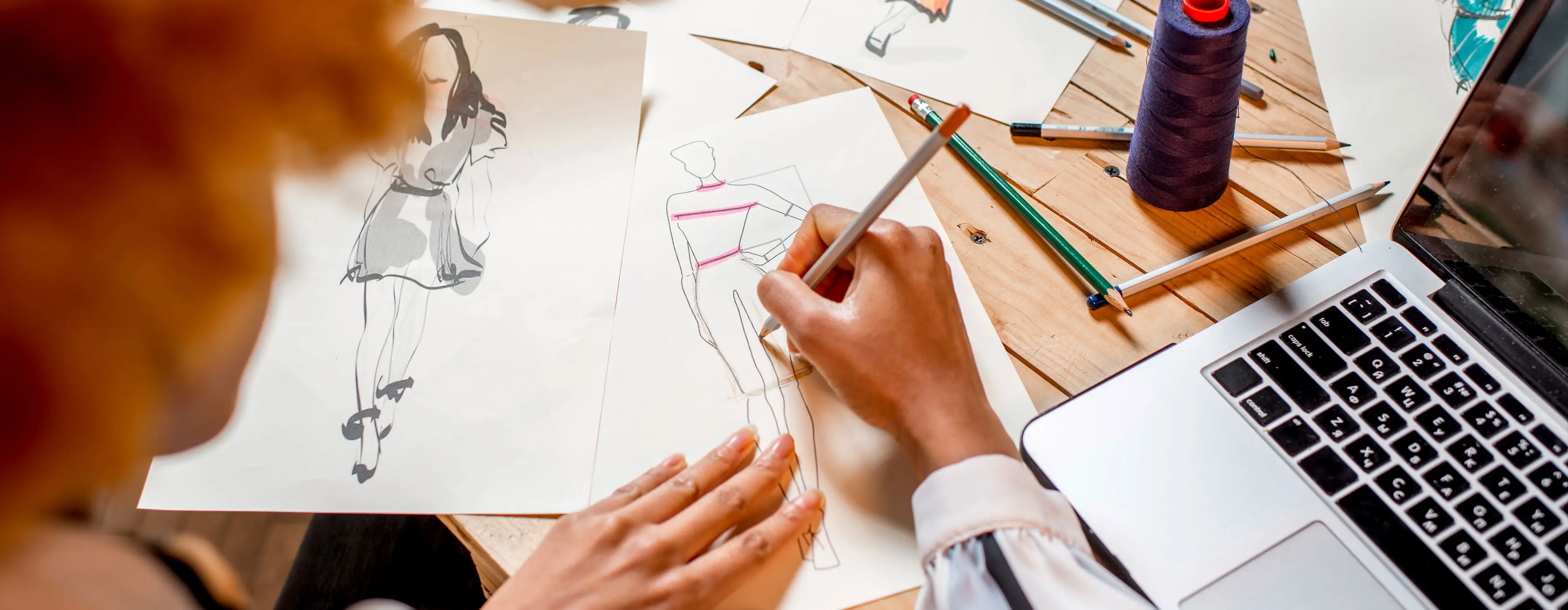 Foto em close das mãos de uma pessoa fazendo um desenho de moda no papel. No cenário, há um computador e mais papéis com outros desenhos.