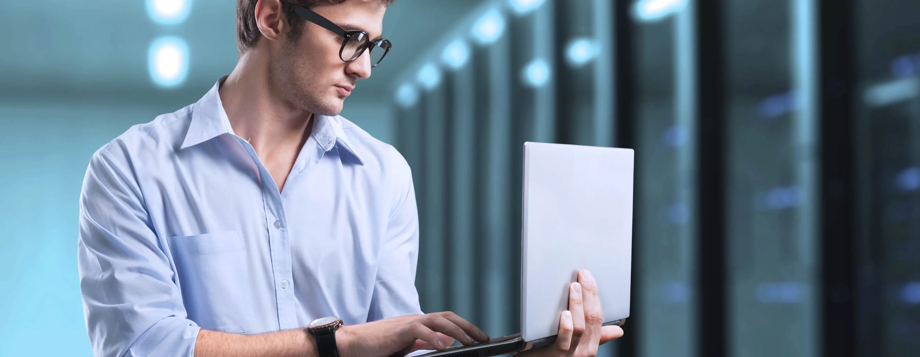 Foto de um homem teclando em notebook de pé em sala de servidores de tecnologia de uma empresa.