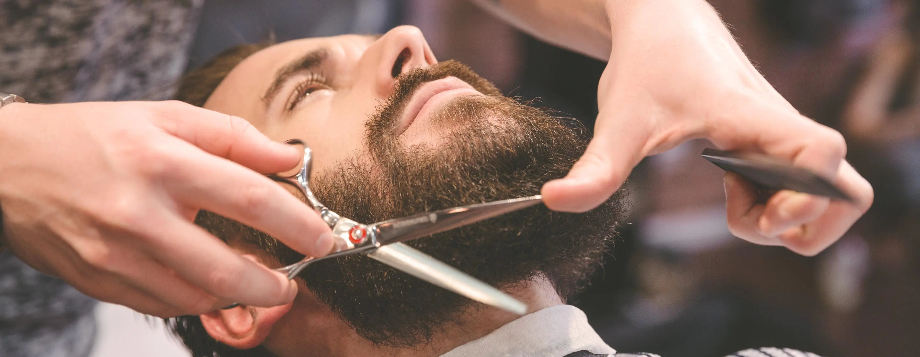 Foto em close de mãos segurando uma tesoura e cortando a barba de um homem.