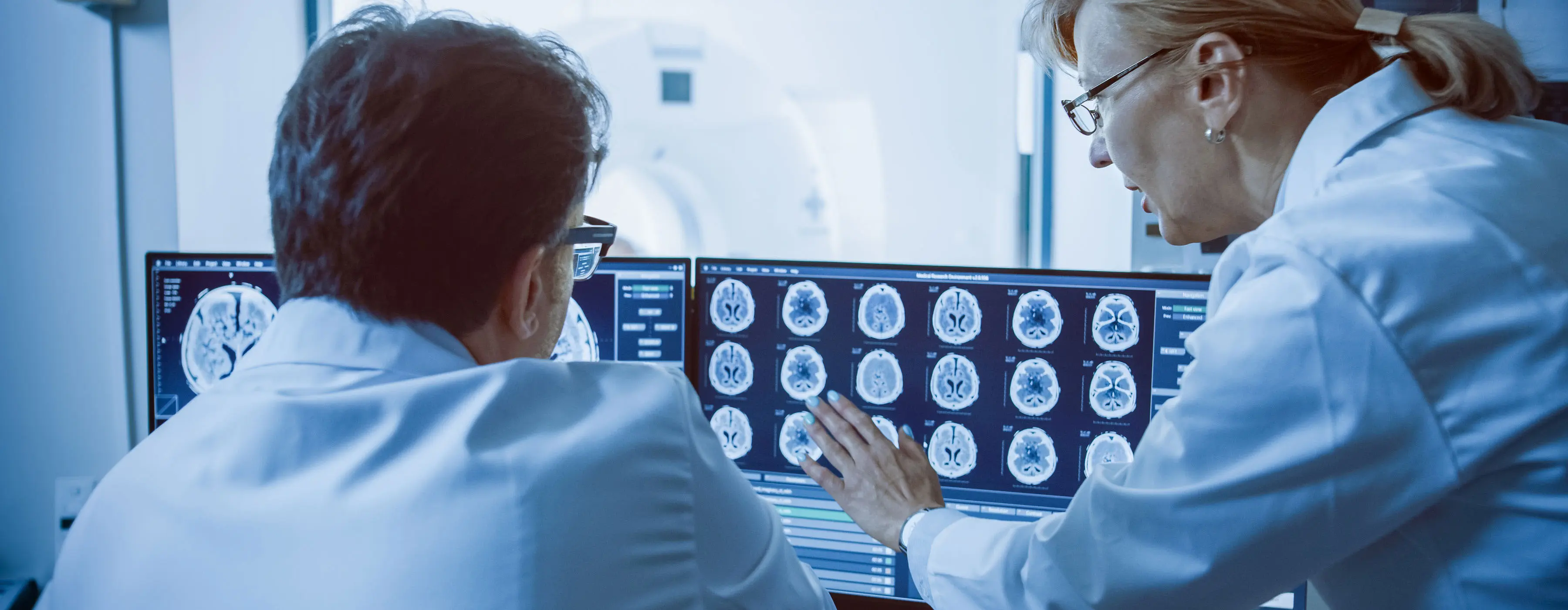 Foto de sala de controle em que dois profissionais de saúde discutem as imagens de varreduras cerebrais. Em segundo plano, o paciente é submetido a procedimento de ressonância magnética ou tomografia computadorizada.
