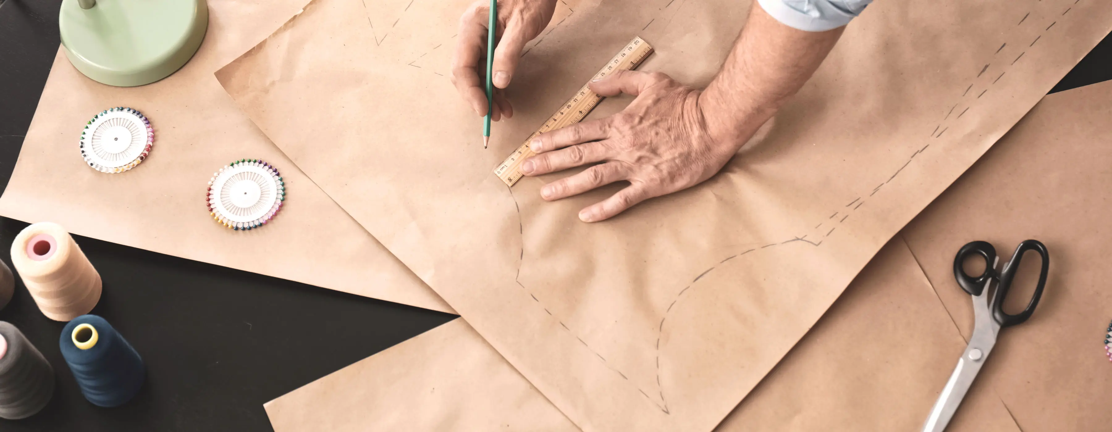 Foto de mãos de uma pessoa desenhando um molde de roupa sobre uma mesa com utensílios de costura.
