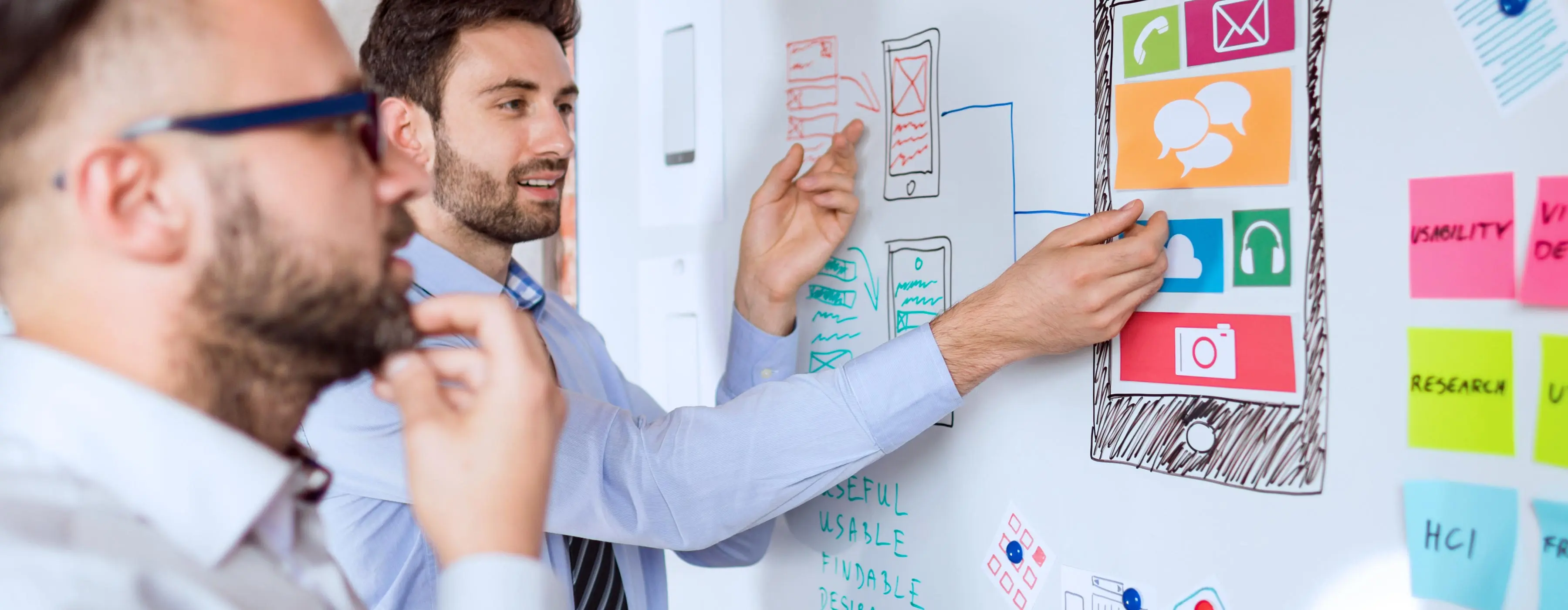 Foto de dois homens conversando e apontando esquema gráfico de projeto digital em um quadro branco.
