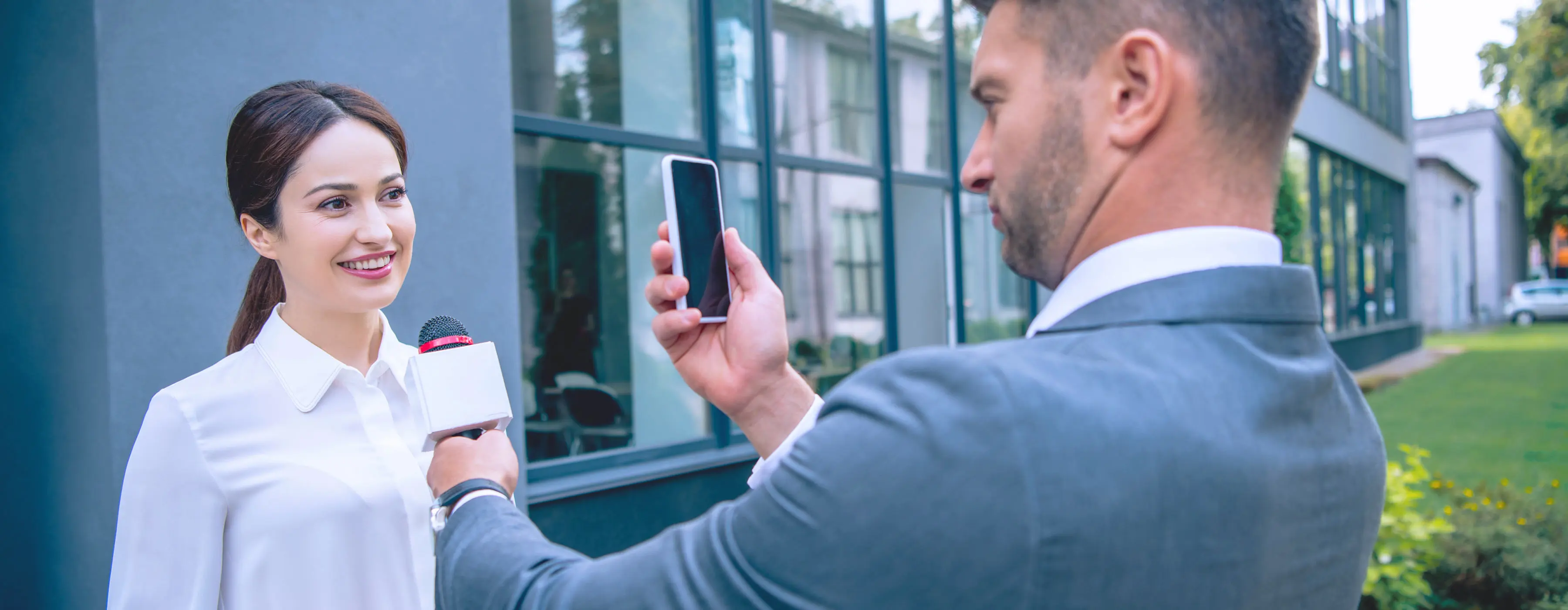 Foto de um homem segurando microfone e celular entrevistando uma mulher na parte externa de um prédio comercial.