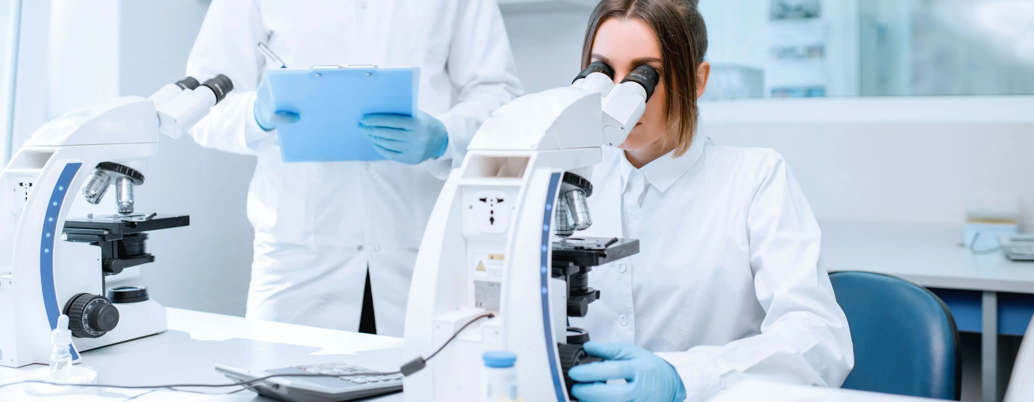 Foto de mulher de jaleco e luvas cirúrgicas, usando um telescópio em um cenário de laboratório.