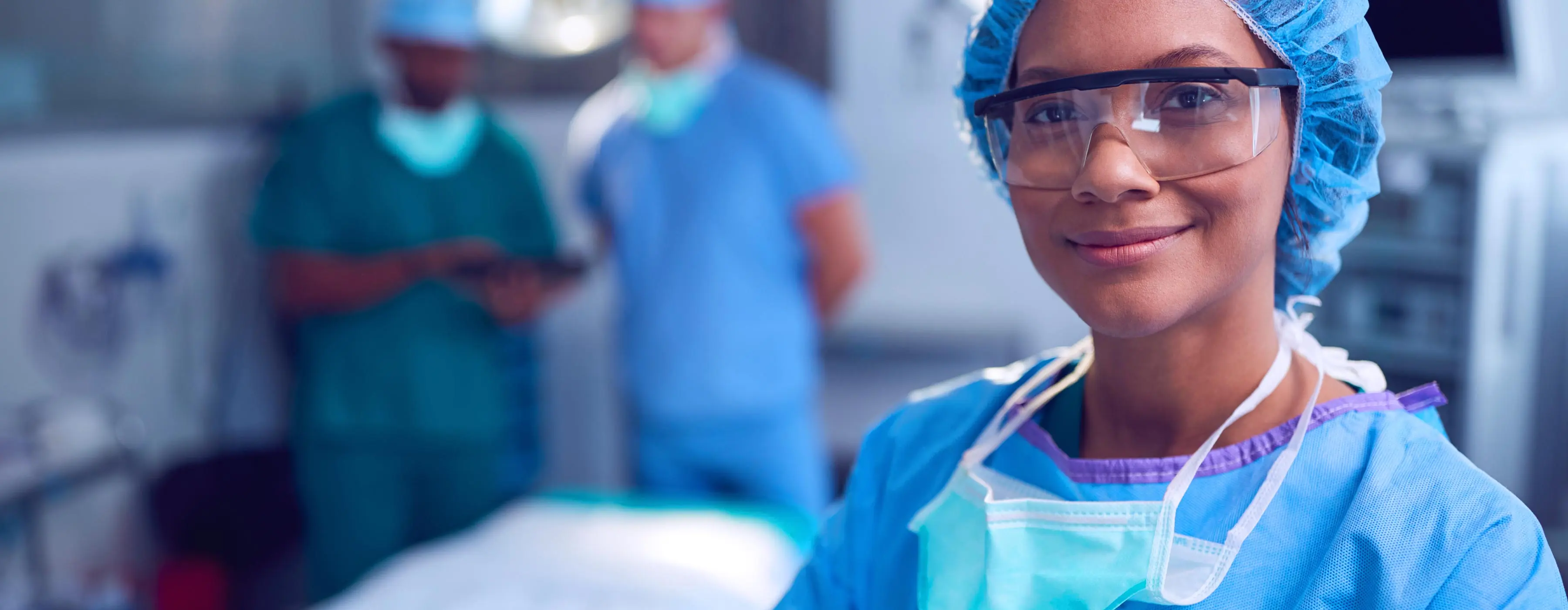 Foto de uma profissional de saúde em ambiente hospitalar, vestindo touca, óculos e avental, com dois colegas de trabalho ao fundo.