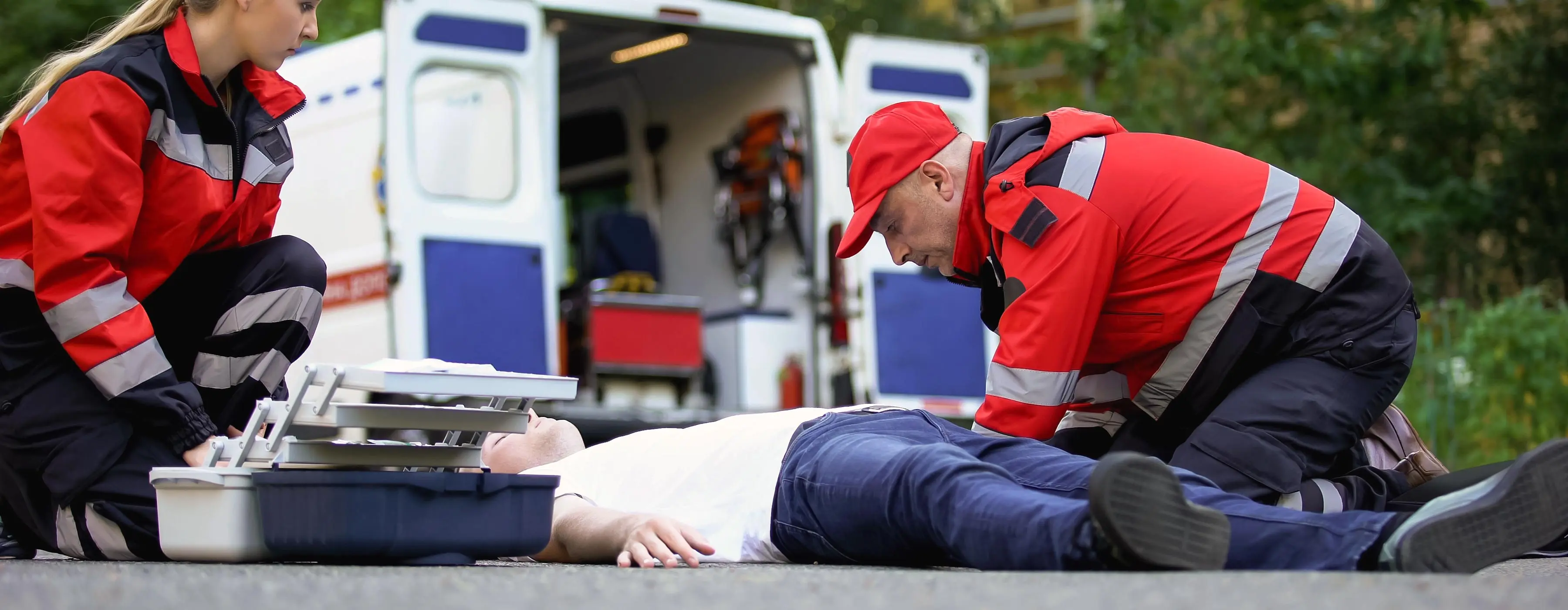 Foto de dois profissionais da saúde ajoelhados no asfalto, em via pública, prestando primeiros socorros a um homem deitado no asfalto, com uma ambulância ao fundo.