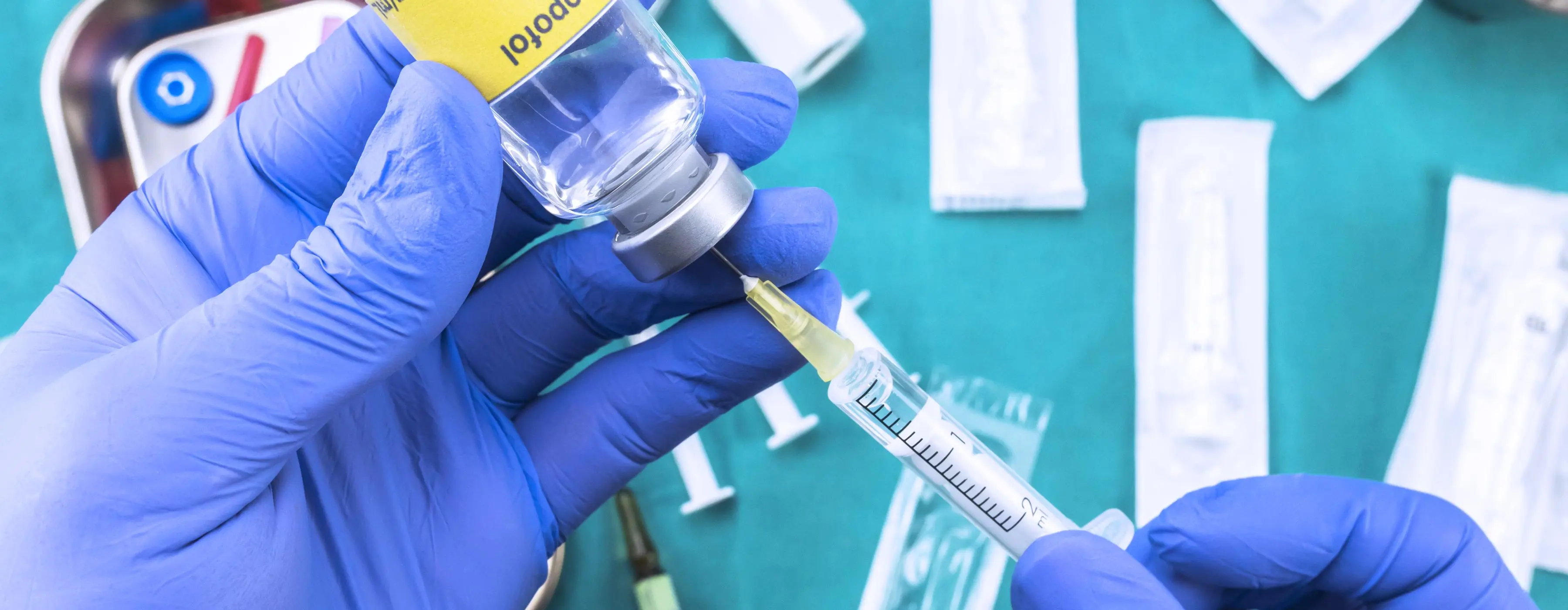 Foto de close de uma pessoa com luvas cirúrgicas usando uma seringa para extrair medicamento de uma ampola e ao fundo mesa com itens hospitalares.