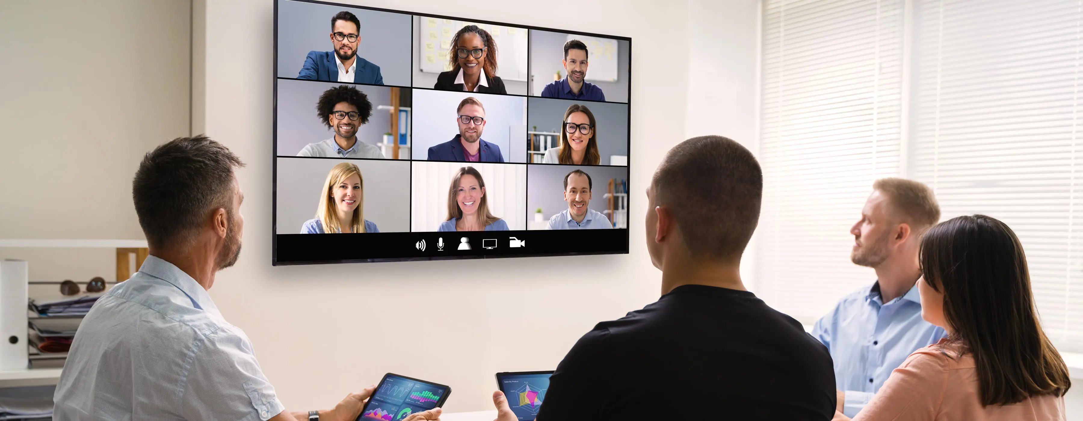 Foto de um grupo de pessoas sentadas em um escritório assistindo em um grande monitor um evento virtual aonde aparecem outros participantes.