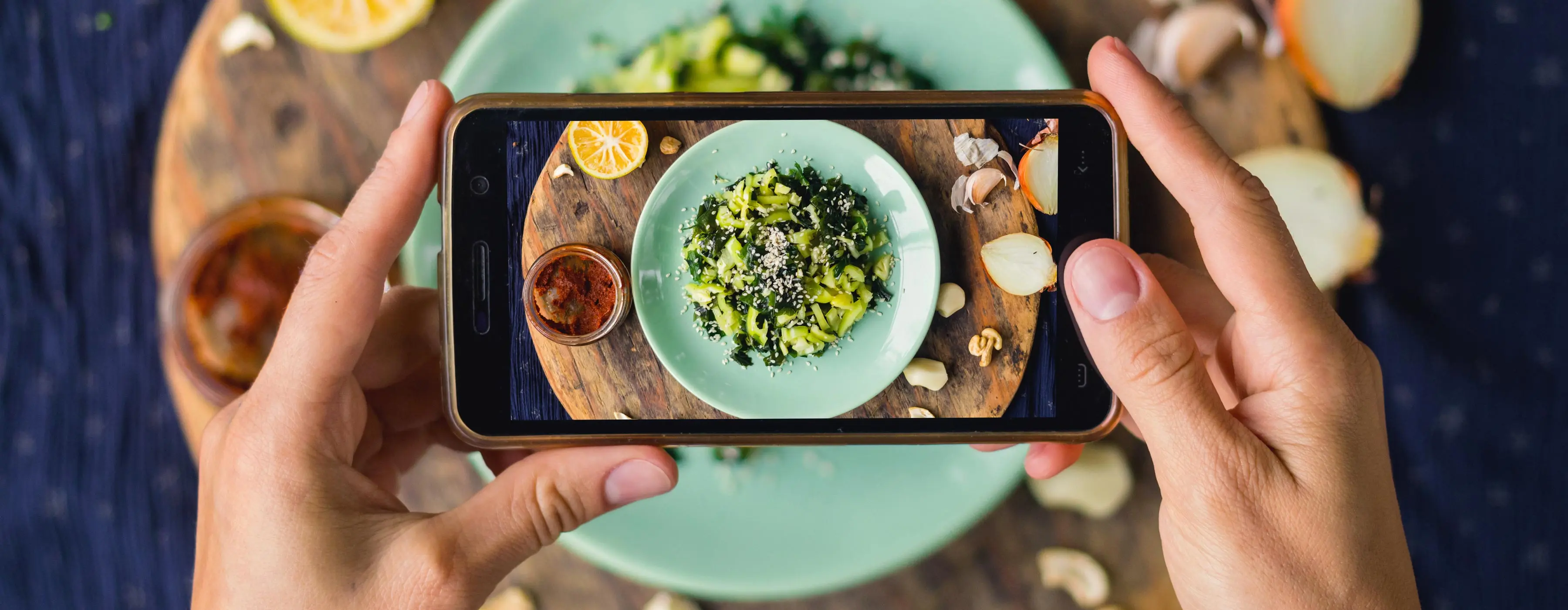 Foto do close das mãos de uma pessoa tirando foto em celular de prato de legumes refogados em um prato sob tábua de madeira com temperos, limão, alho e cebola.