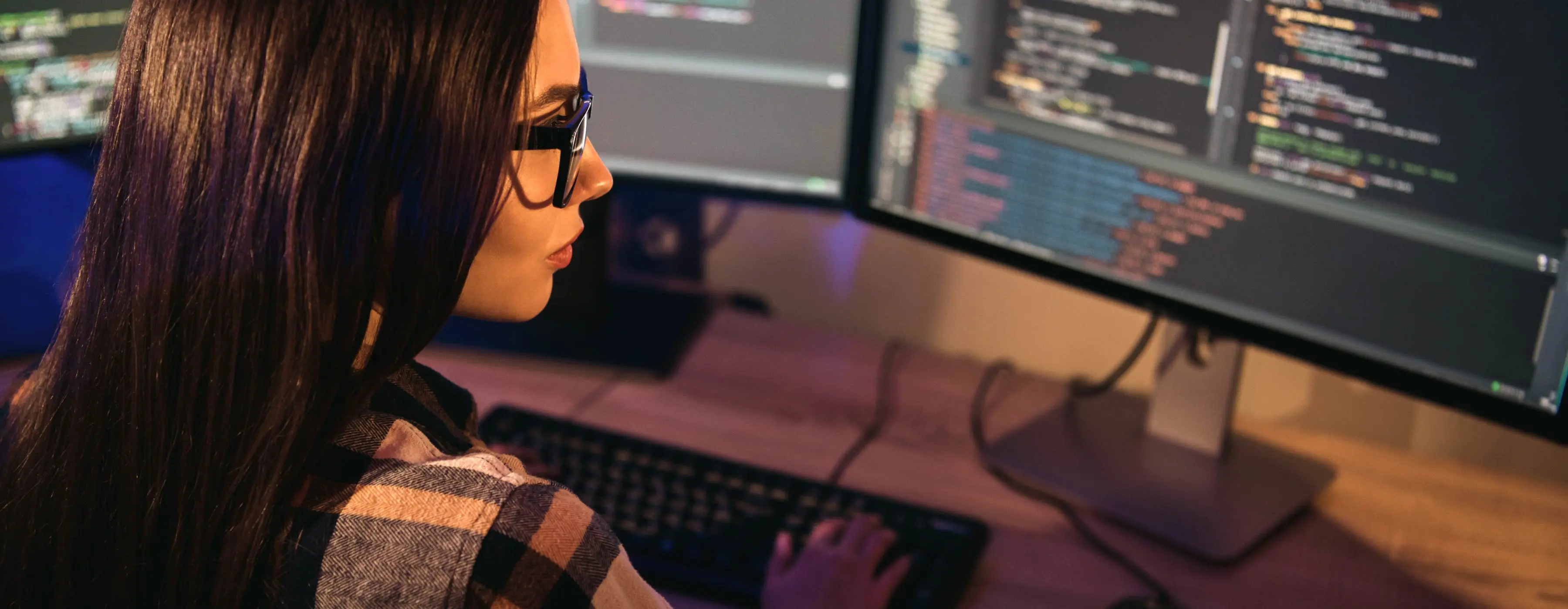 Foto de jovem digita dados observando três telas de computador em escritório.