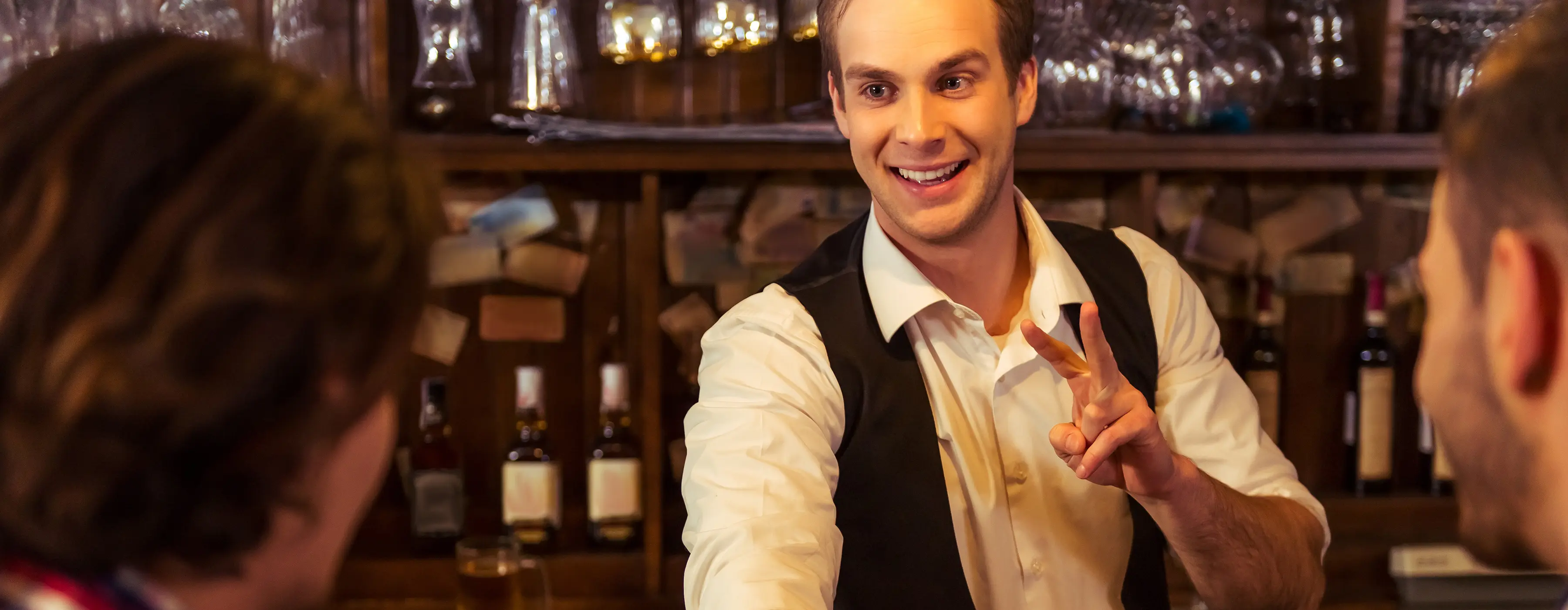 Foto de homem bartender sorrindo com dois dedos levantado enquanto atende duas pessoas