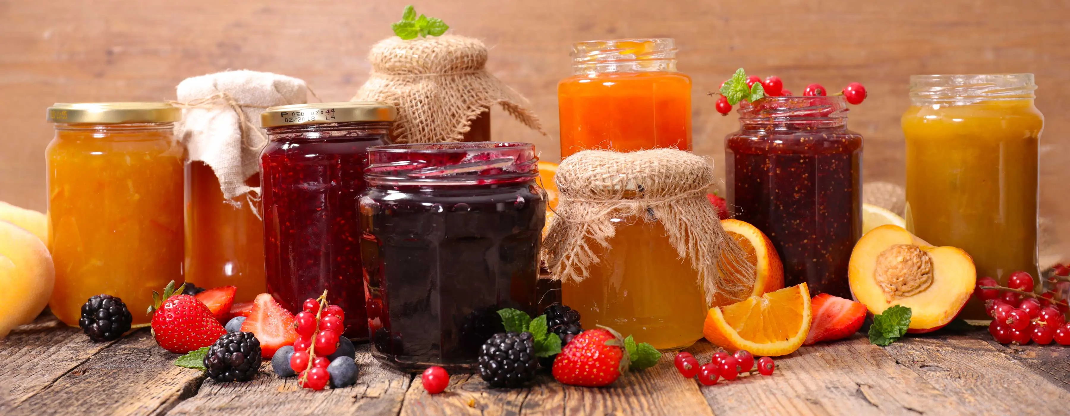 Foto de mesa com frutas espalhadas (morangos, amoras e cerejas) e diversos potes de geleias frescas prontas para o consumo.