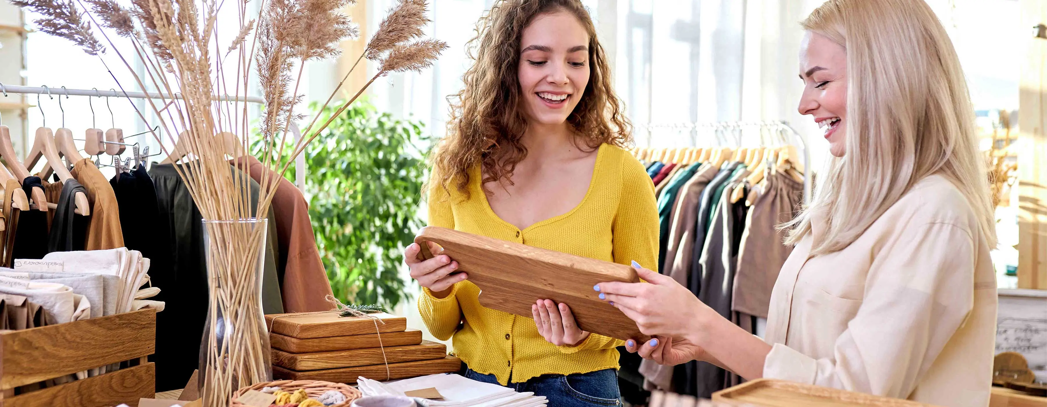 Foto de duas mulheres sorridentes em ambiente com roupas e acessórios ao redor, segurando tábua de madeira entre as mãos.