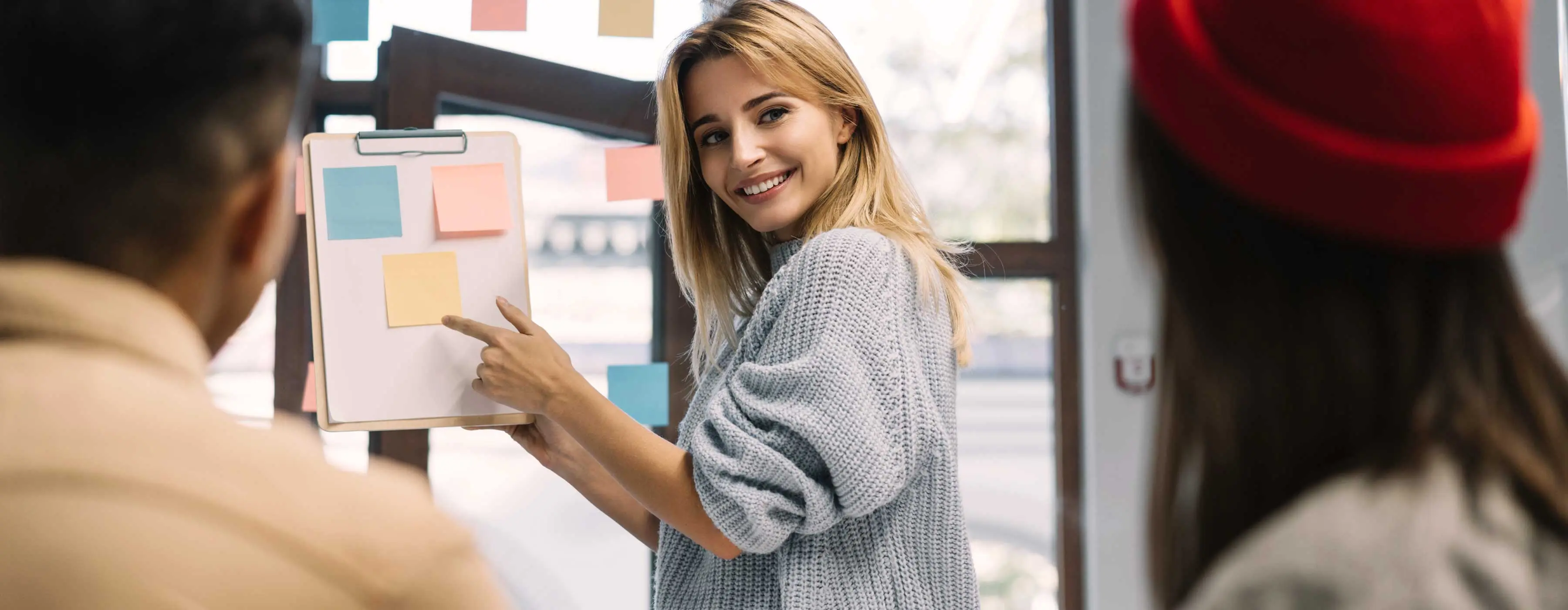 Foto de mulher sorrindo para outras pessoas enquanto segura prancheta com notas adesivas coloridas entre as mãos.