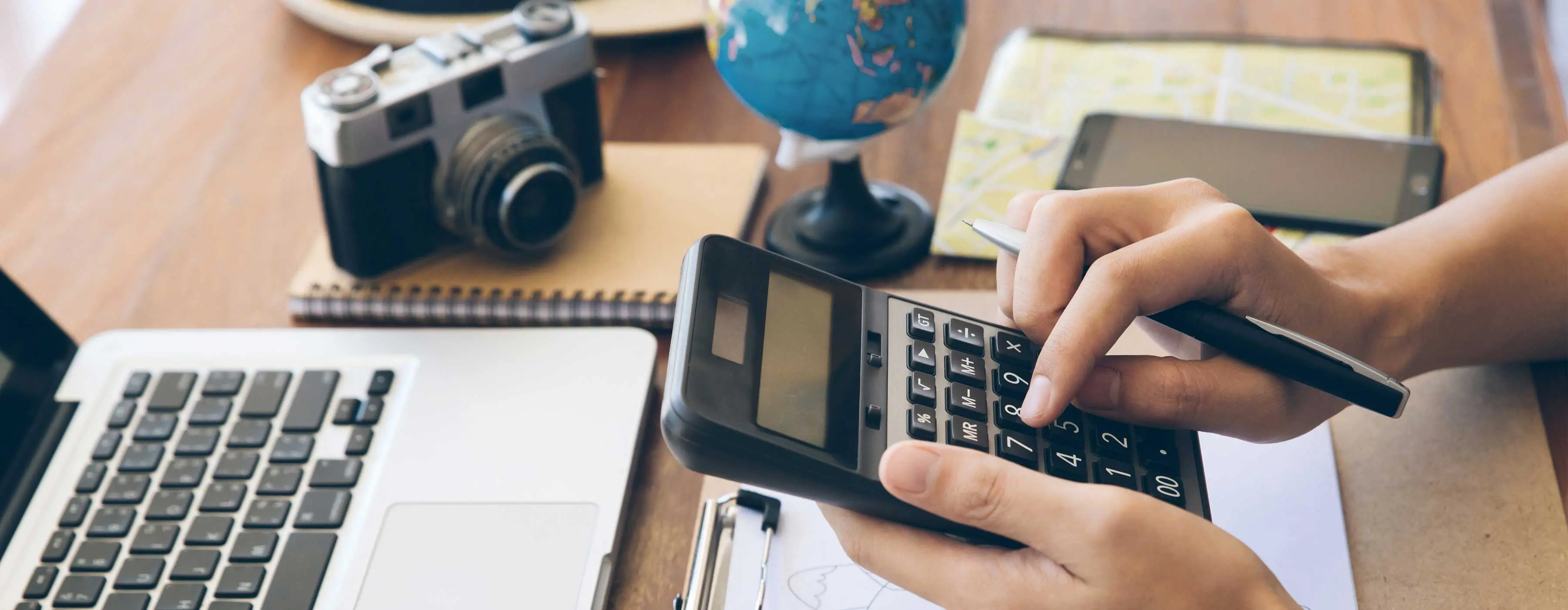 Foto focando as mãos de uma pessoa usando uma calculadora, sentado em mesa com notebook, máquina fotográfica, blocos de anotação, prancheta com desenho e notas de dólar. 
