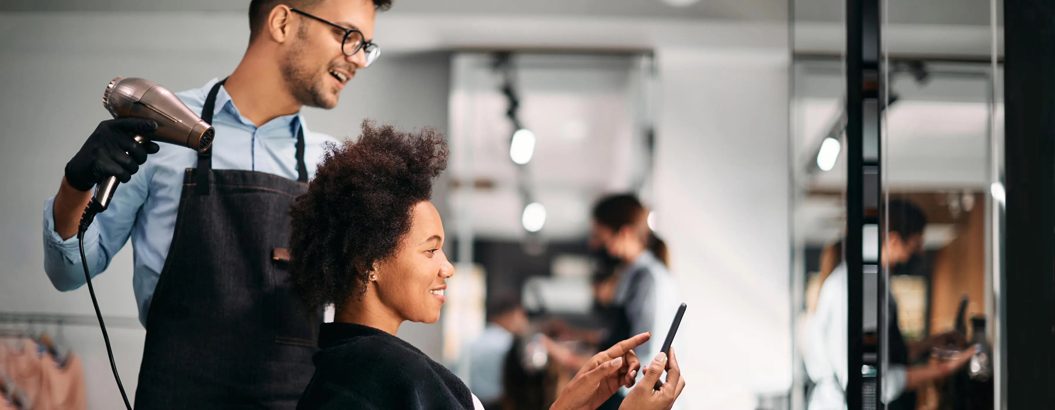 Foto de um profissional em um salão de beleza usando luvas, segurando um secador de cabelo e atendendo moça de cabelos crespos que mostra algo para ele na tela do celular