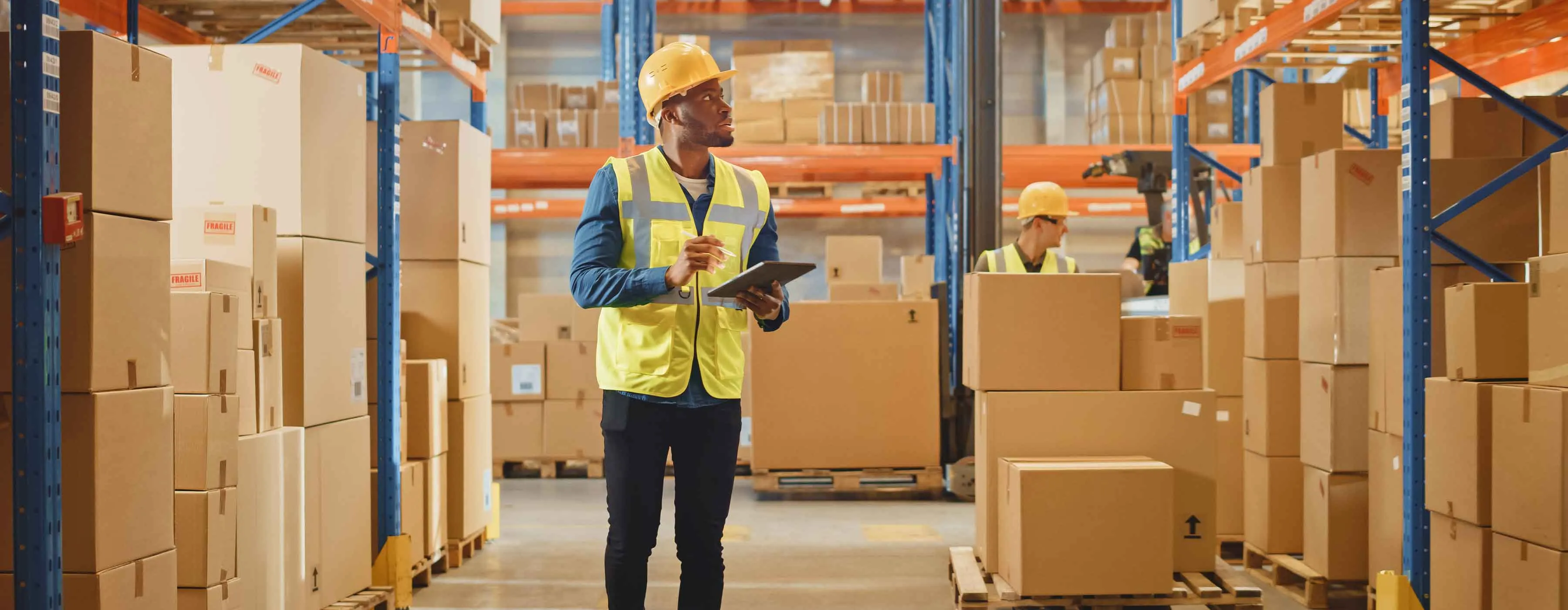 Homem vestindo equipamentos de segurança verifica estoque de empresa anotando dados em uma prancheta. 