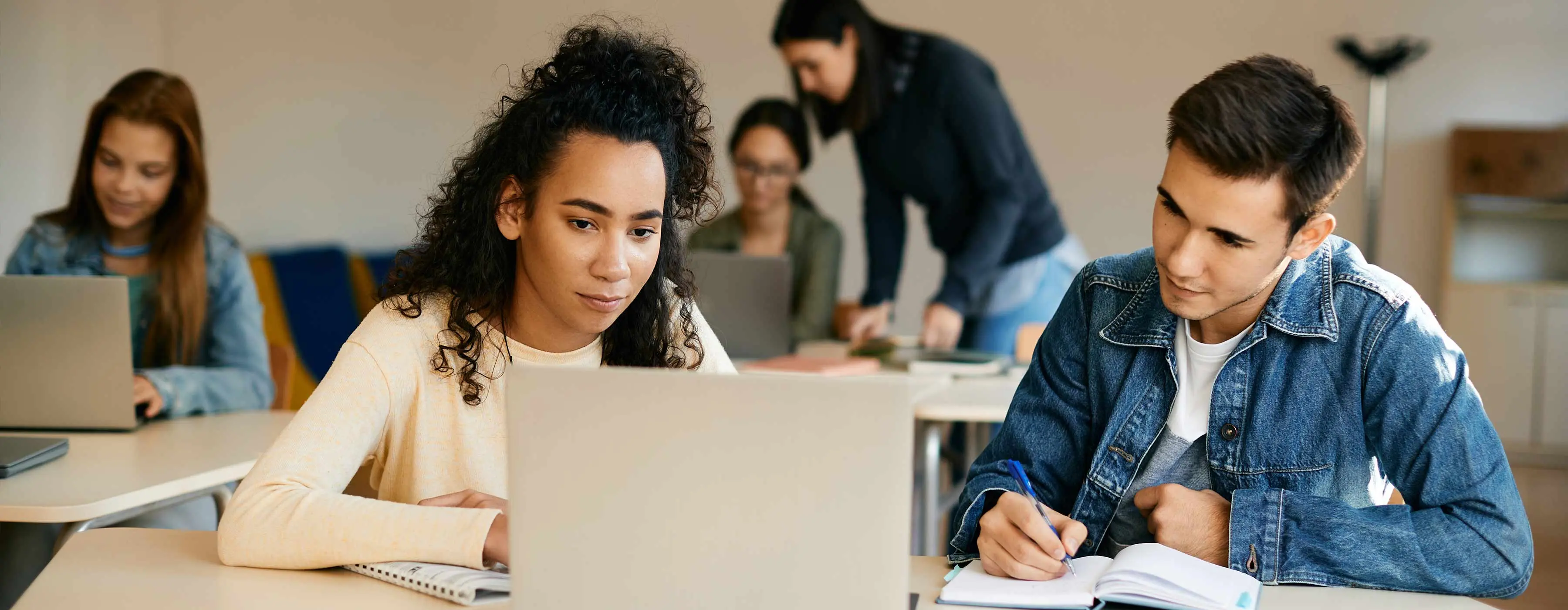 Foto de duas pessoas sentadas, olhando para um notebook e fazendo anotações. O cenário é de uma sala com outras pessoas fazendo o mesmo ao fundo.