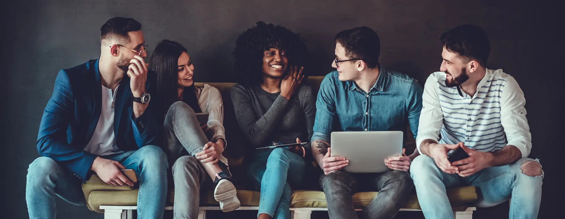 Foto de grupo de moças e rapazes, sentados ao lado um do outro, em sala comum, sorrindo e conversando.