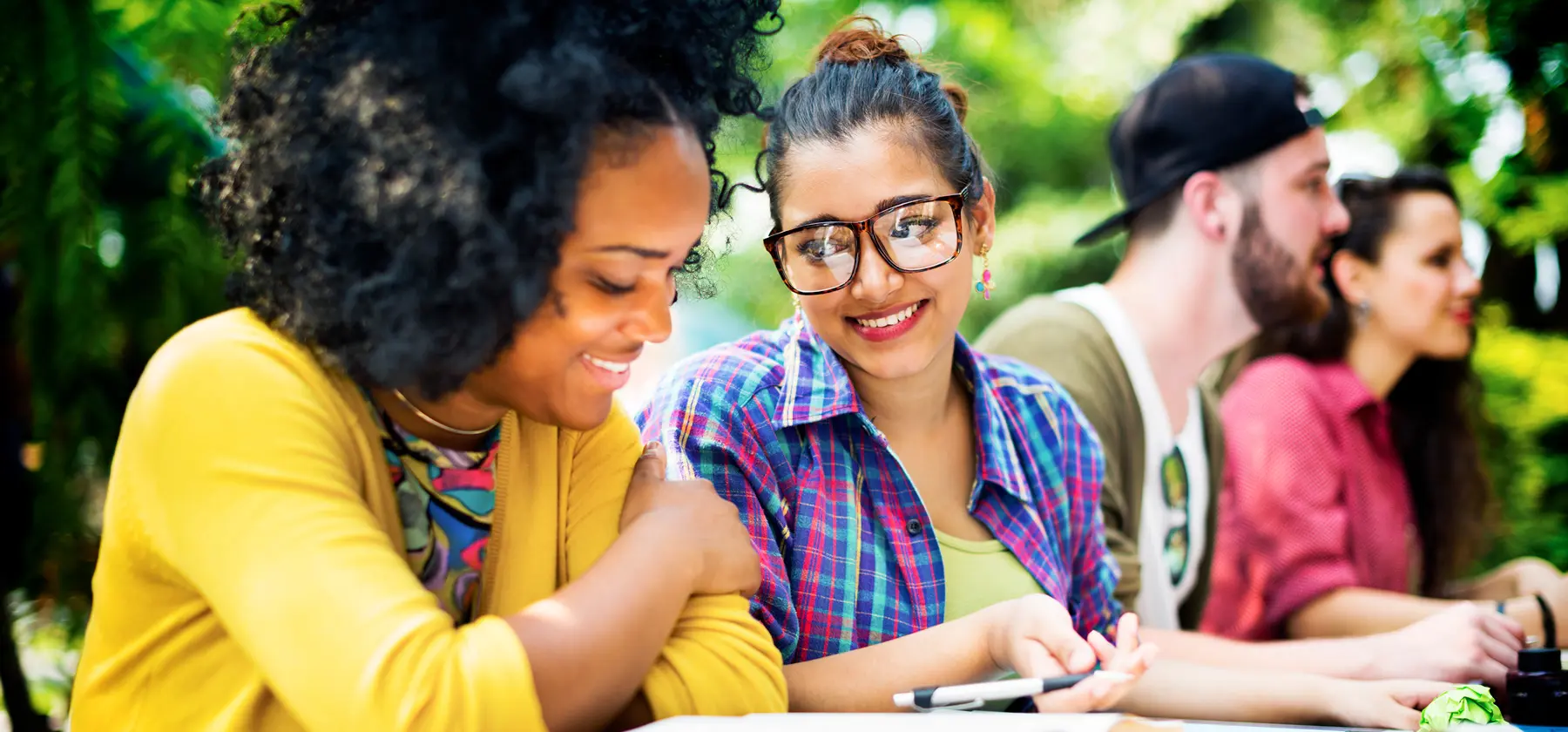 Foto de duas mulheres jovens sorrindo e sentadas lado a lado ao ar livre, com folhagens e pessoas ao fundo.