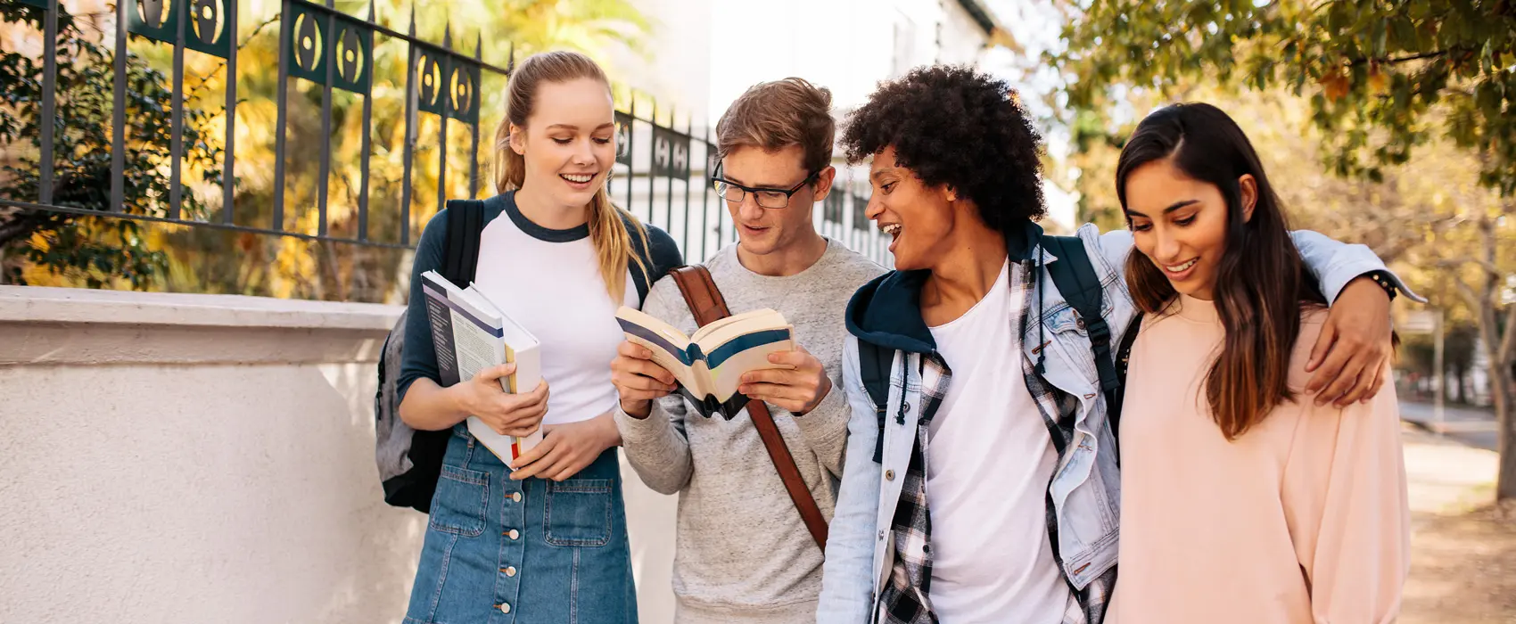 Foto de quatro jovens lado a lado sorrindo em calçada arborizada. Um dos jovens segura um livro aberto entre as mãos.