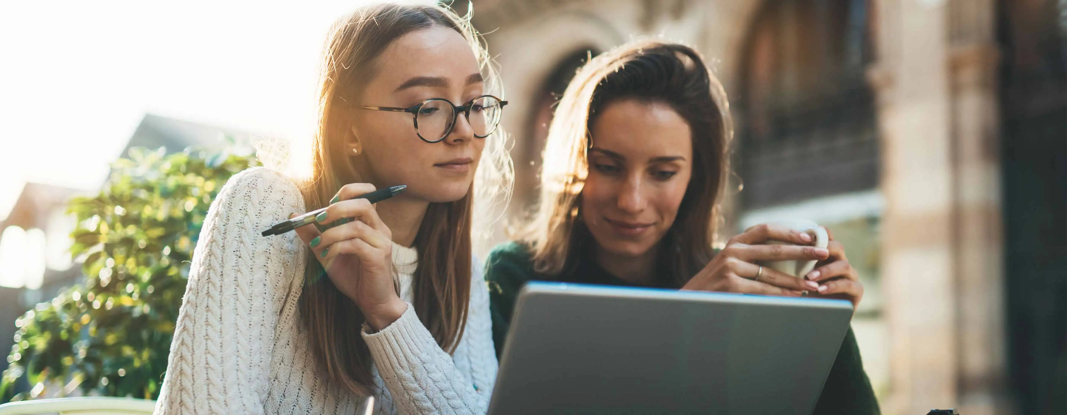 Foto de duas moças sentadas ao ar livre olhando para a tela do notebook.