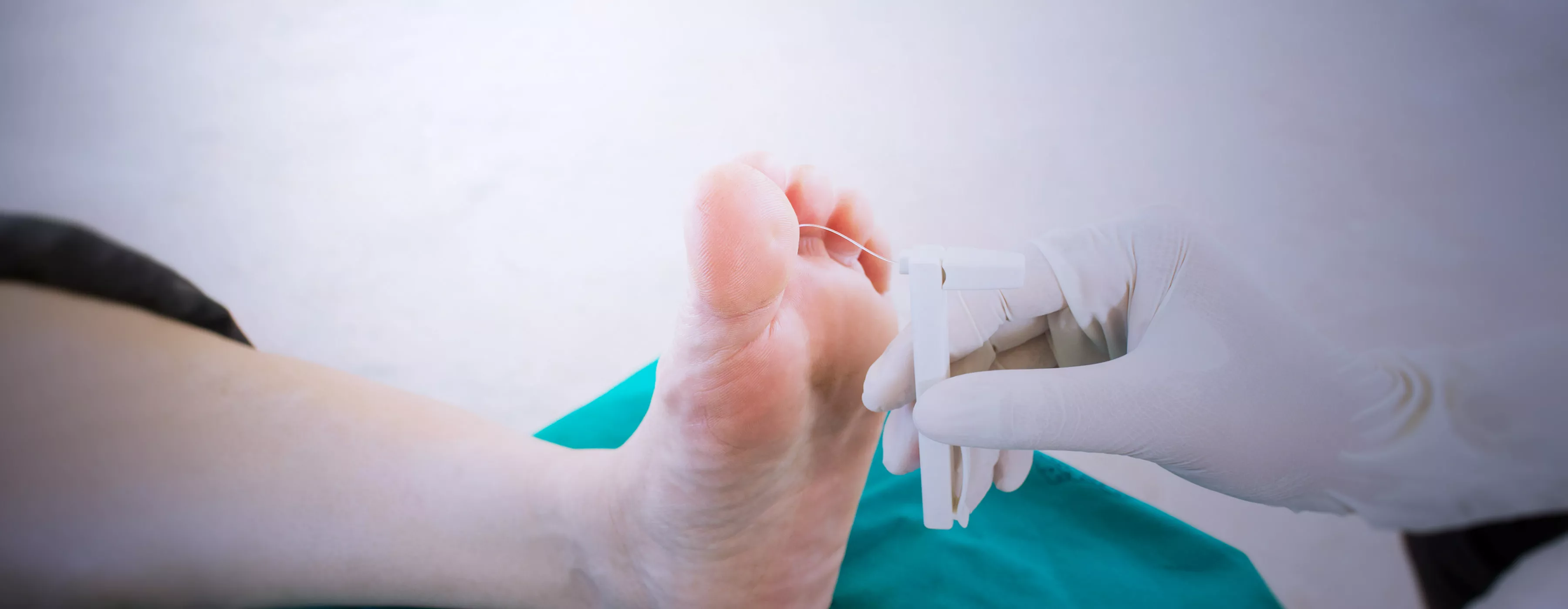 Foto em close de mão de profissional com luva cirúrgica usando um aparelho entre os dedos de um dos pés de outra pessoa.