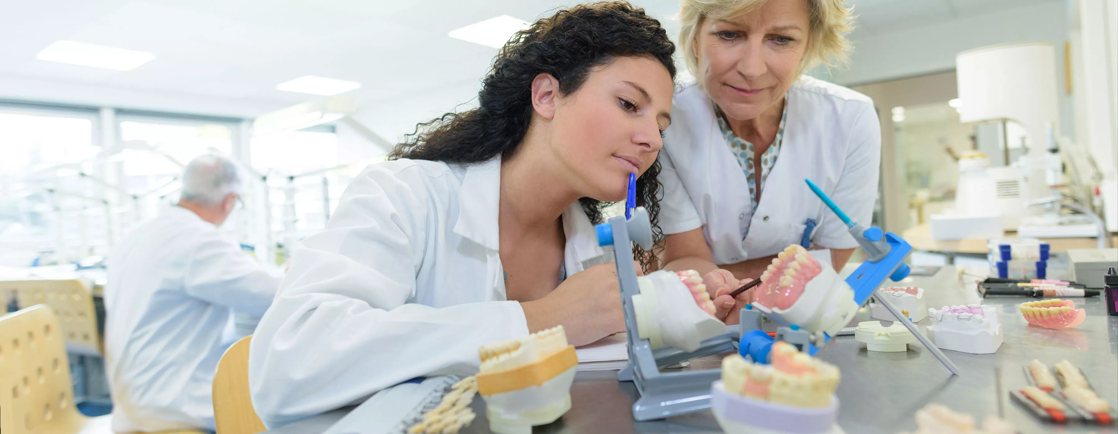 Foto de duas mulheres com aventais observando arcadas dentárias. As duas vestem aventais e estão em laboratório de clínica odontológica.