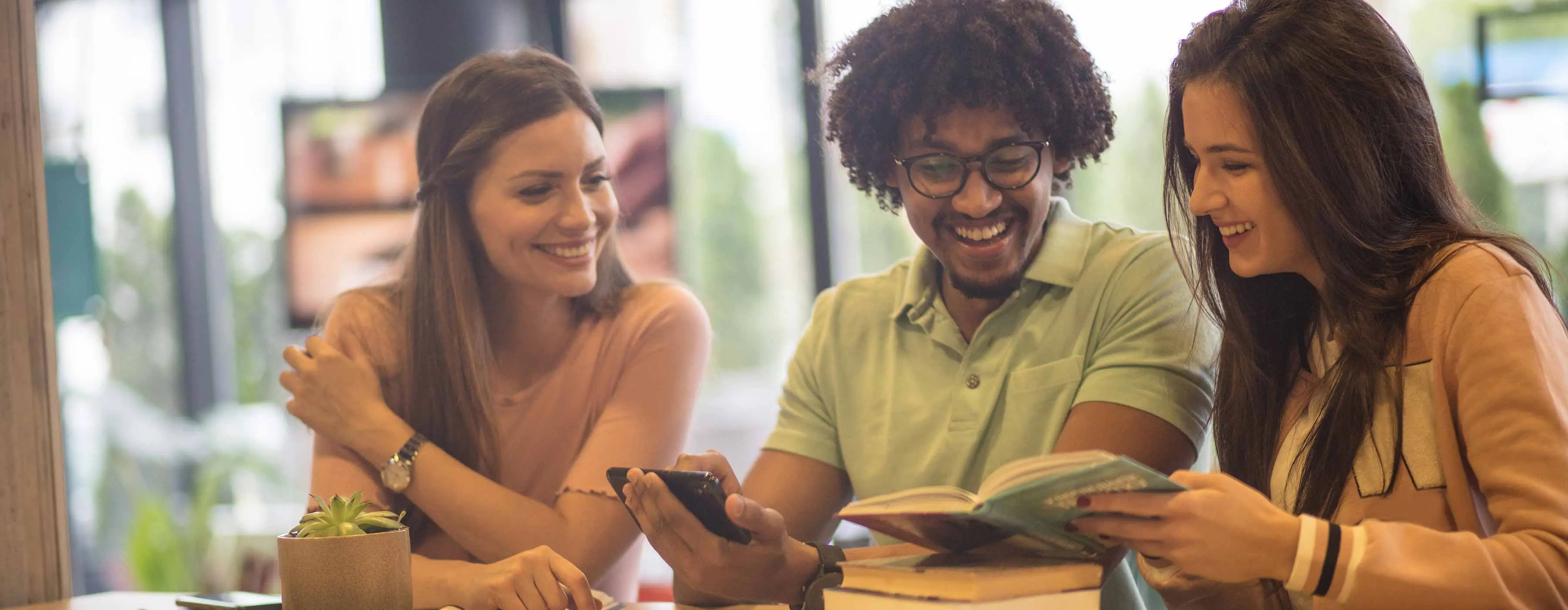 Foto de duas moças e um homem que sorriem enquanto olham para um livro. O homem segura um celular