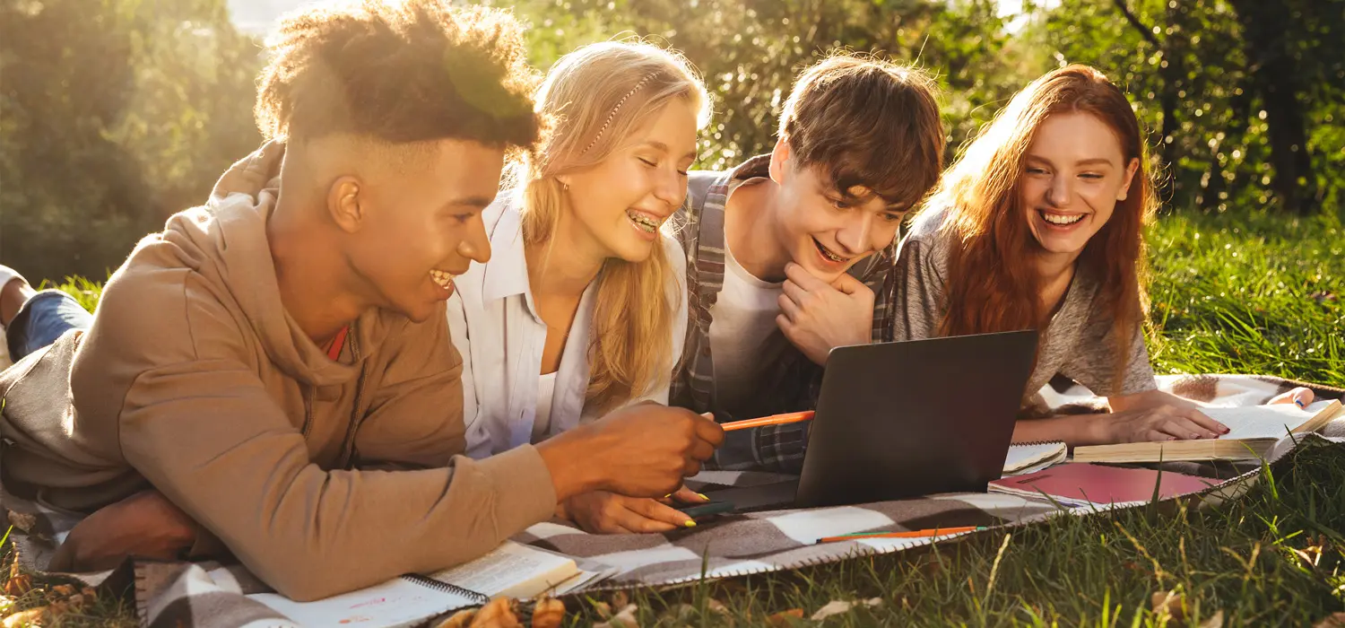 Foto de quatro jovens deitados de barriga na grama, sorrindo olhando para um notebook