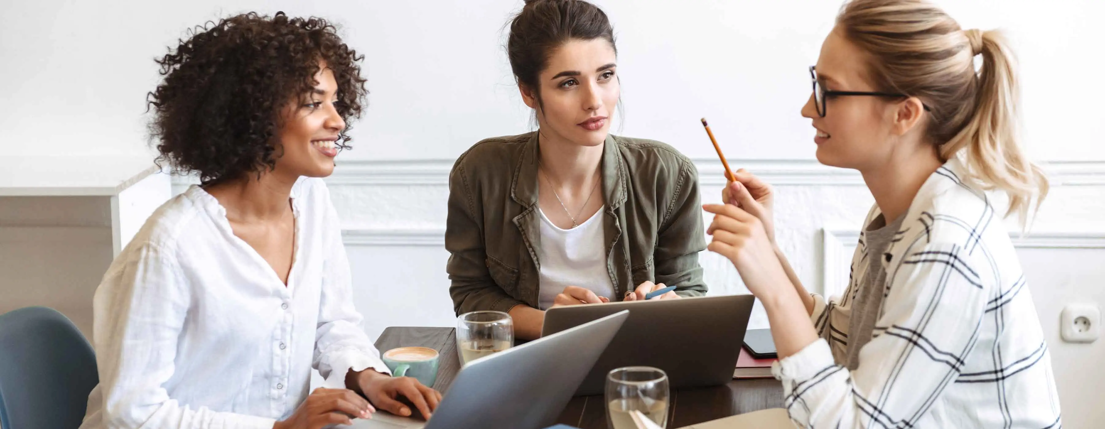 Um grupo de jovens mulheres conversa em uma mesa enquanto usam computadores portáteis. 