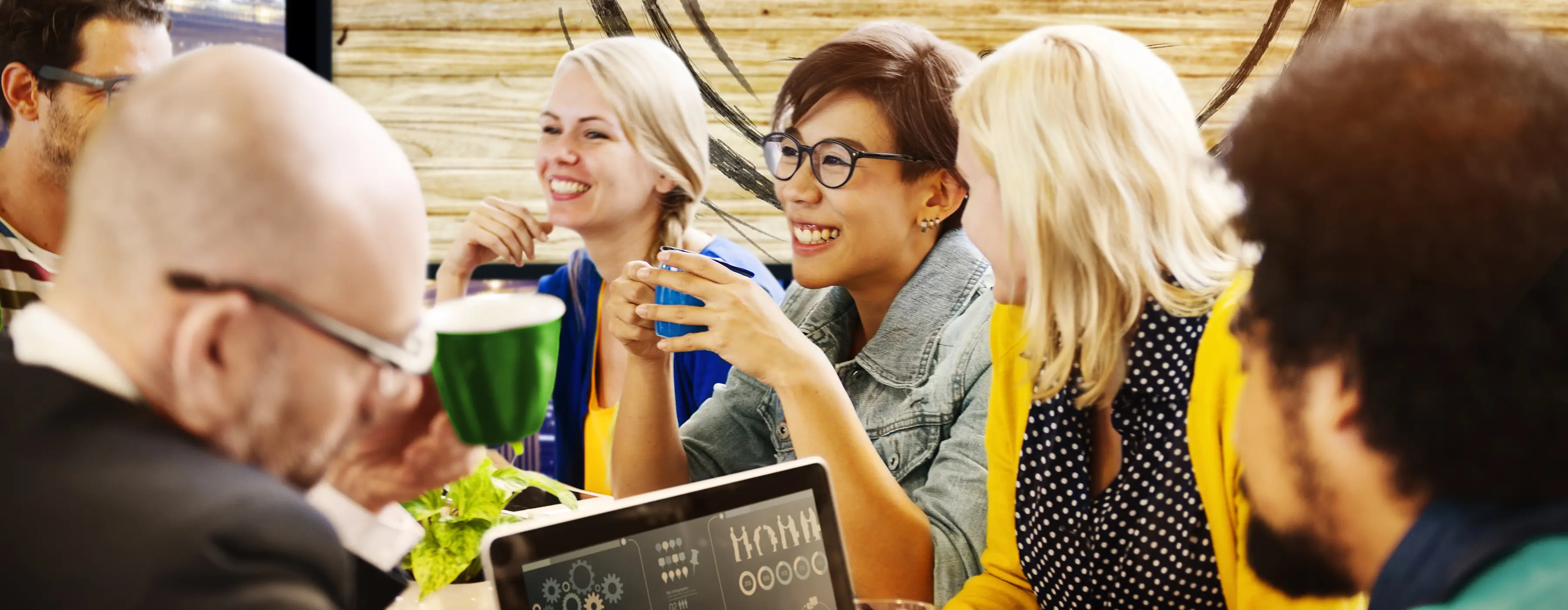 Foto de um grupo de pessoas em uma reunião descontraída, alguns estão segurando uma xícara com bebidas e um outro participando está mexendo em um notebook.