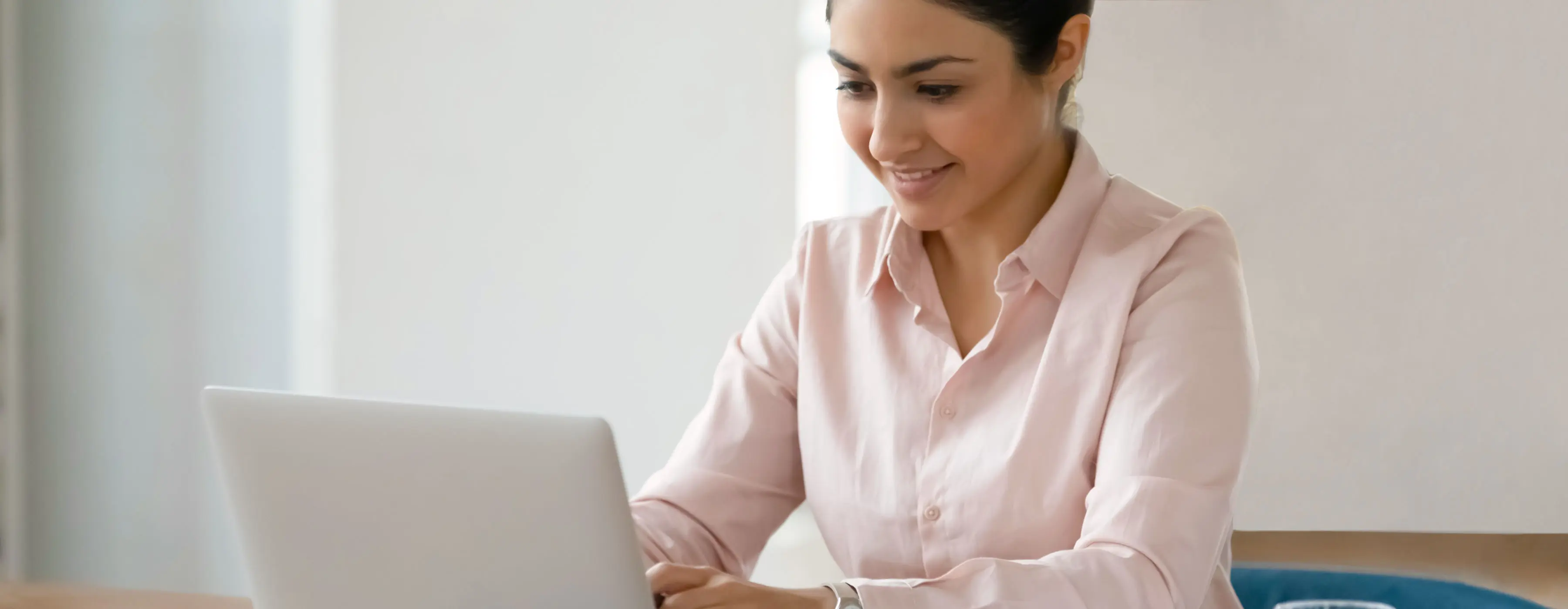 Foto de uma mulher jovem sorridente sentada próxima a uma mesa no local de trabalho digitando em seu notebook e observando atentamente o tela.