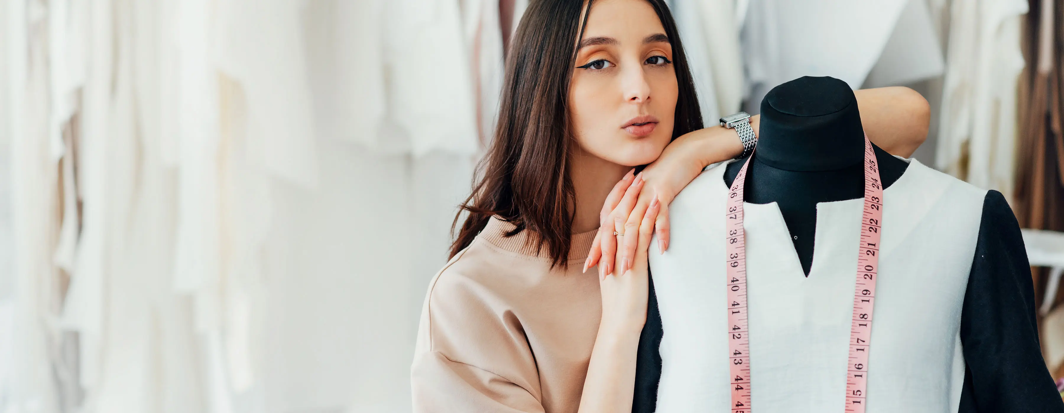 Foto de uma mulher olhando para a câmera, em um ambiente de produção moda, encostada em um manequim que está vestido e com uma fita métrica no pescoço.