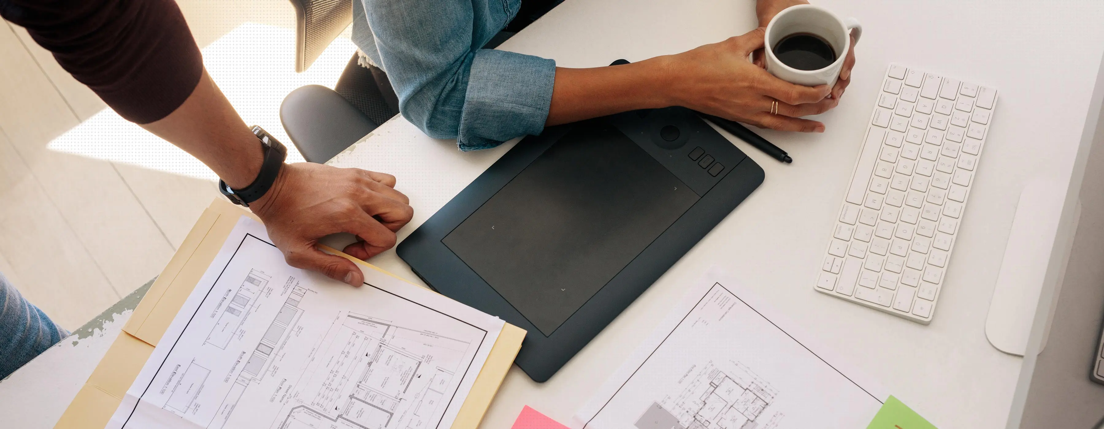 Foto aérea em close das mãos de duas pessoas, em ambiente profissional, verificando projetos arquitetônicos em cima na mesa, que tem um teclaro, xícara de café e equipamento de digitalizar imagem.