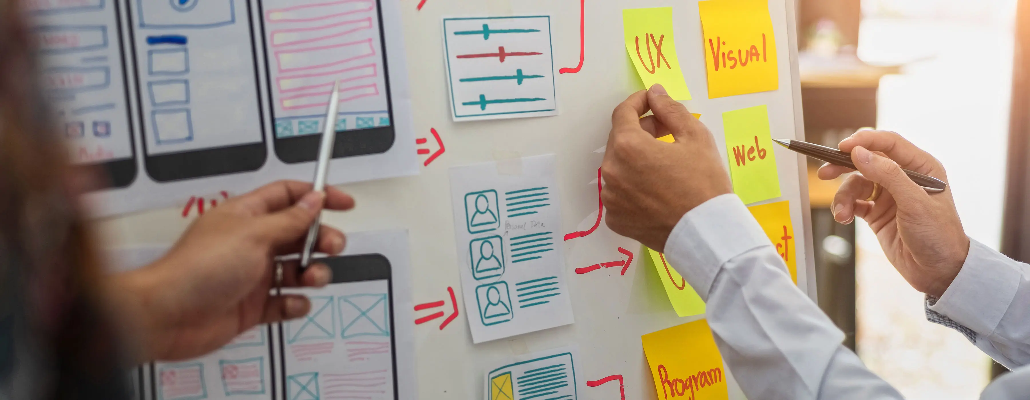 Foto em close de um mural branco onde constam desenhos de protótipos de projetos de programação para aplicativos móveis e diversas anotações em papéis coloridos colados na parede, com planos de desenvolvimento criativo de sites.