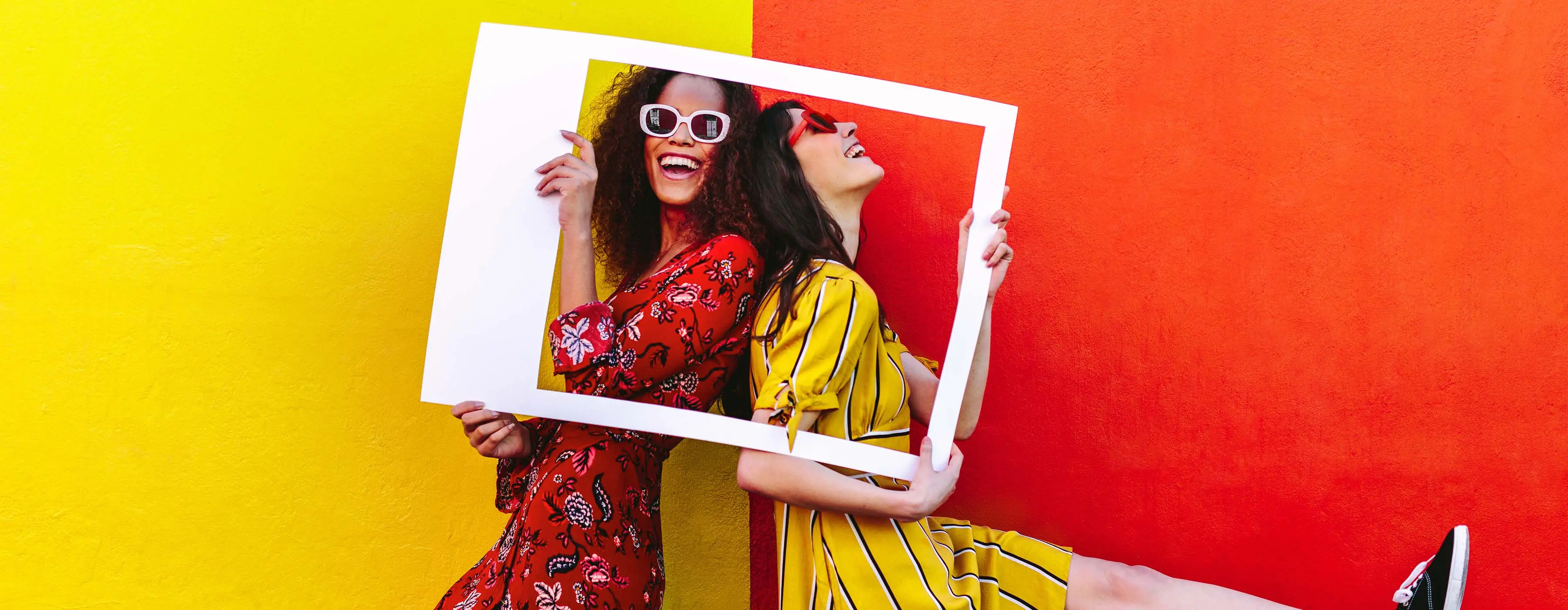 Foto de duas jovens sorrindo posando para editorial de moda, usando vestidos coloridos e óculos escuros.