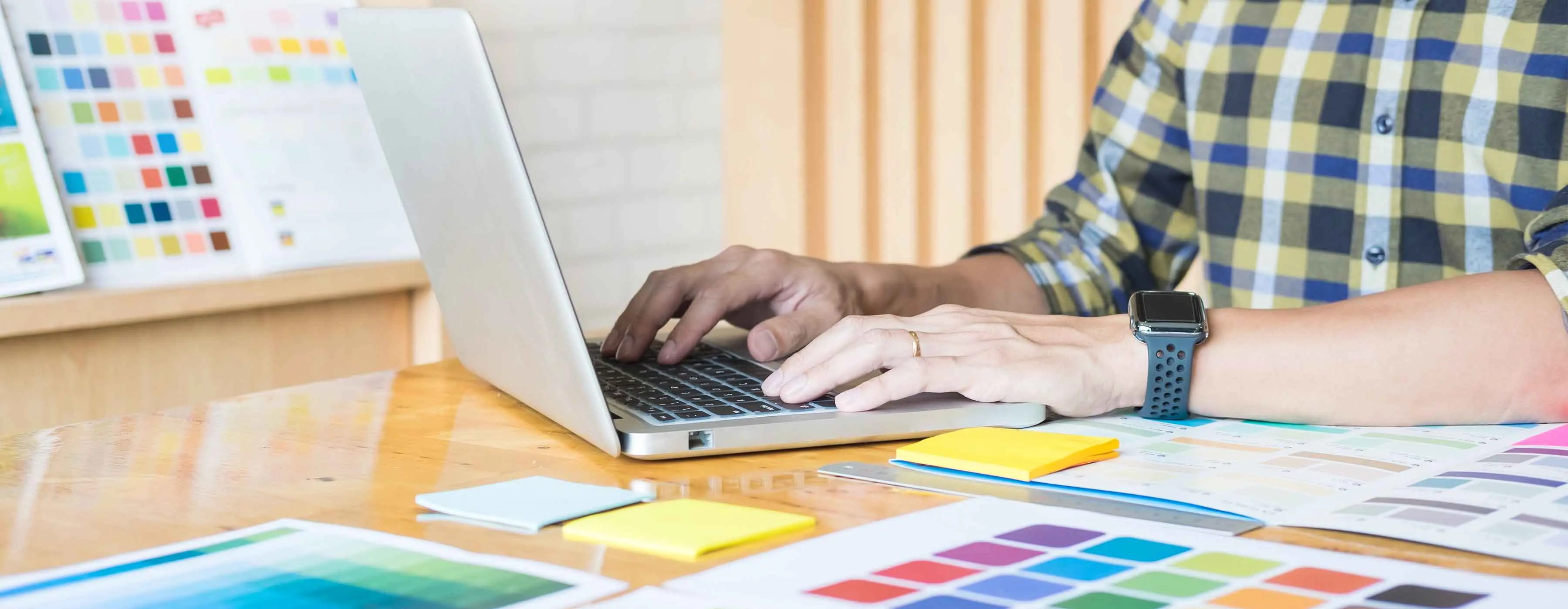 Foto de uma pessoa mexendo no notebook e do lado dela em cima da mesa várias paletas de cores para trabalho de designer gráfico.