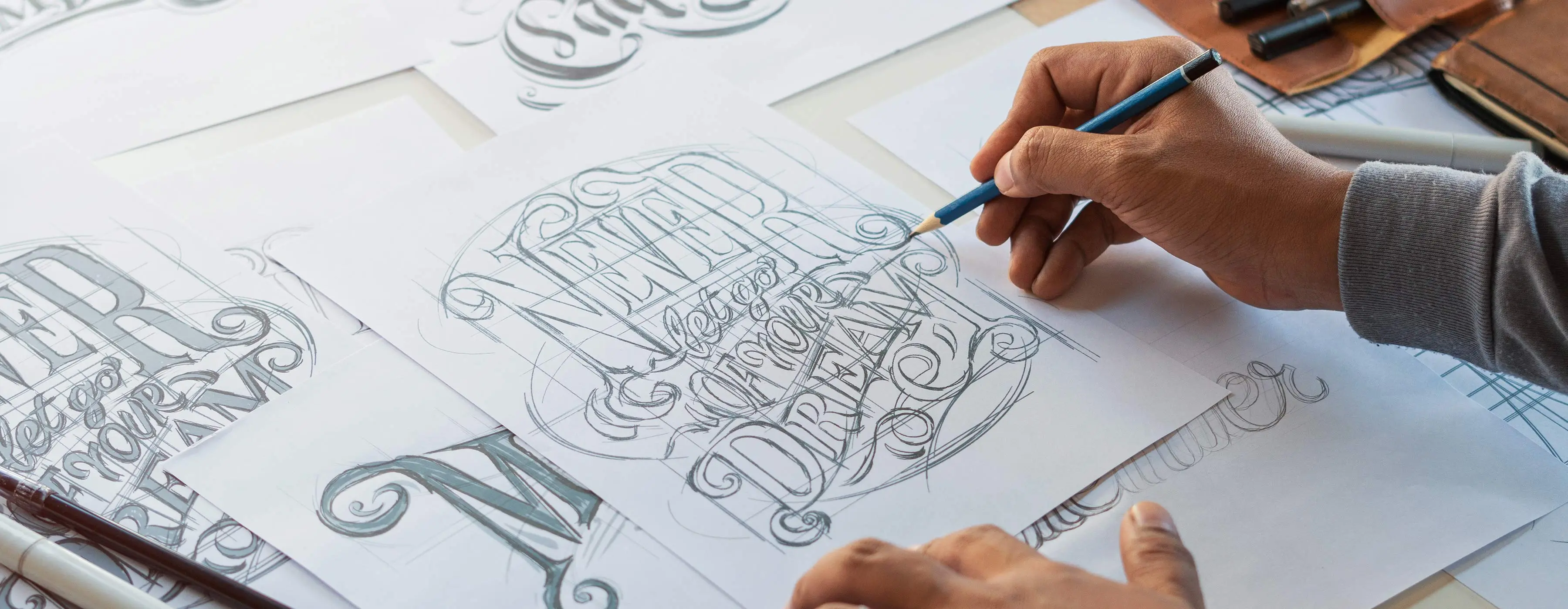 Foto em close de um artista desenhando um esboço tipográfico e algumas letras caligrafadas com pincel e tinta em um estúdio de design.