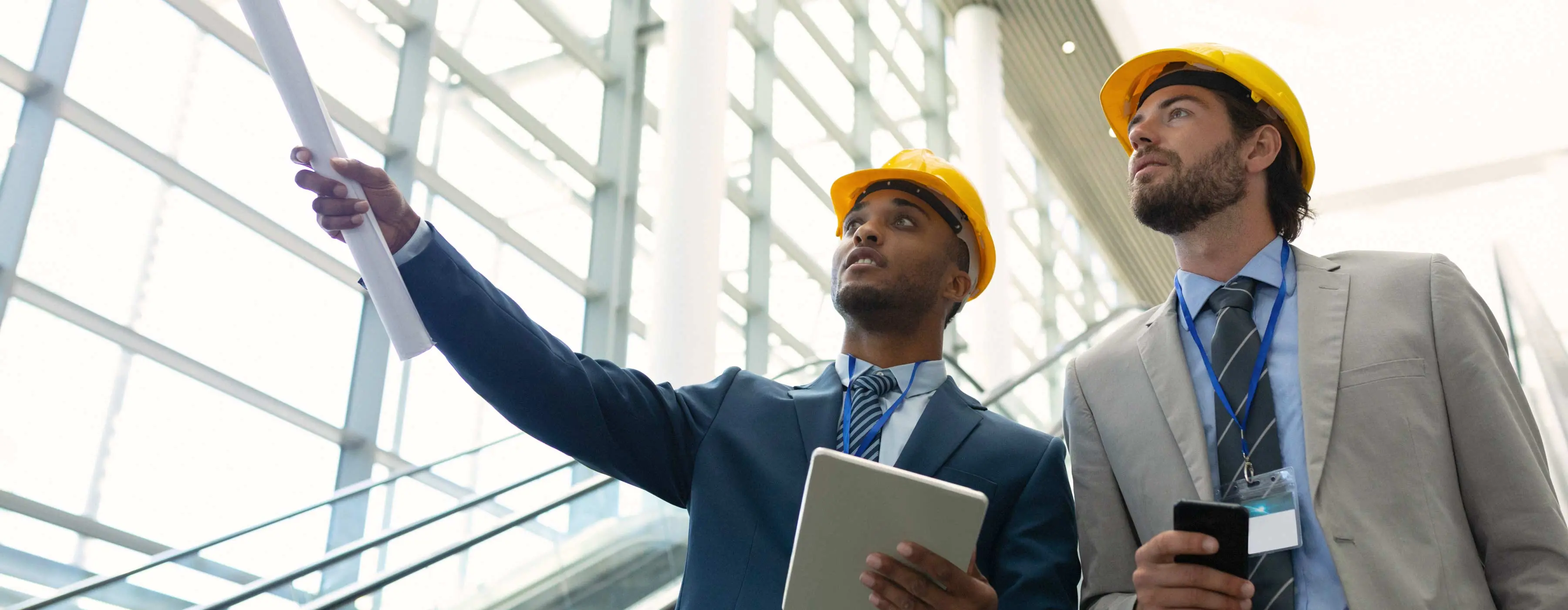 Foto de dois homens com roupas formais e usando capacete, descendo a escada rolante de um prédio comercial e conversando sobre a arquitetura do ambiente.