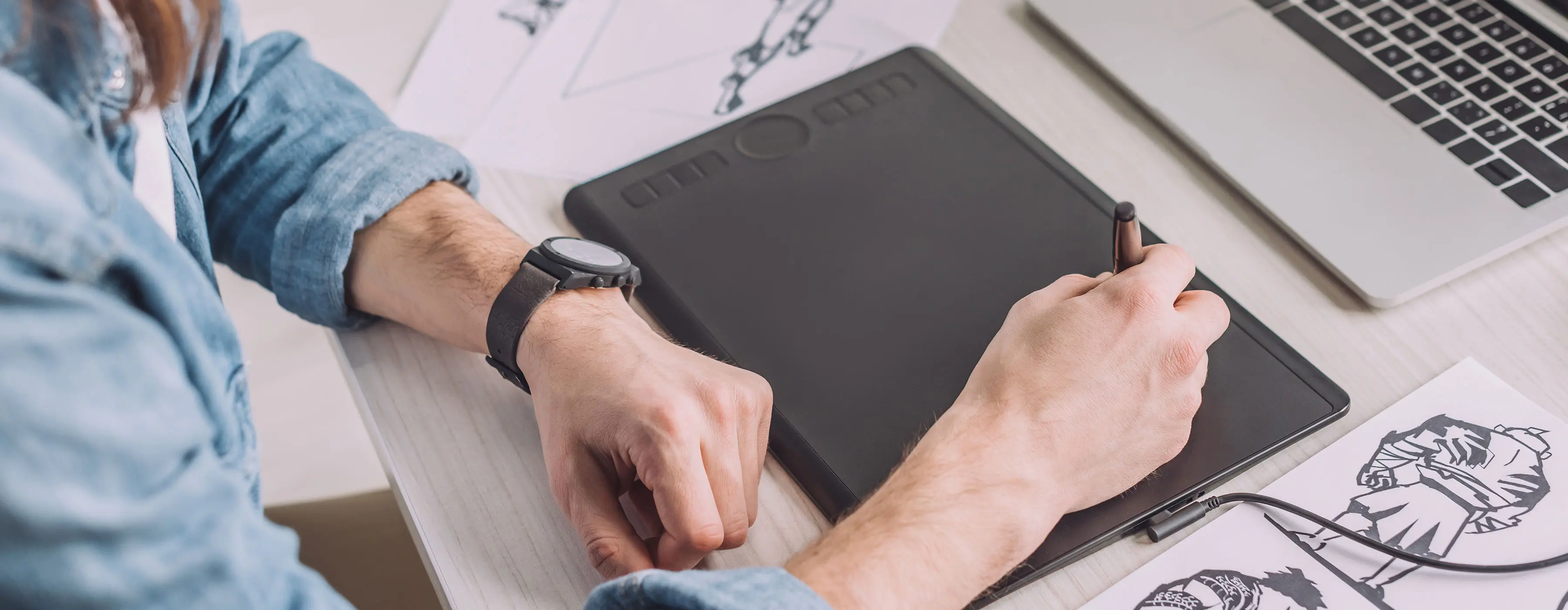 Foto em close que mostra as mãos de uma pessoa desenhando com o uso de uma caneta em um mesa digitalizadora, próxima a alguns desenhos impressos e um teclado de computador.