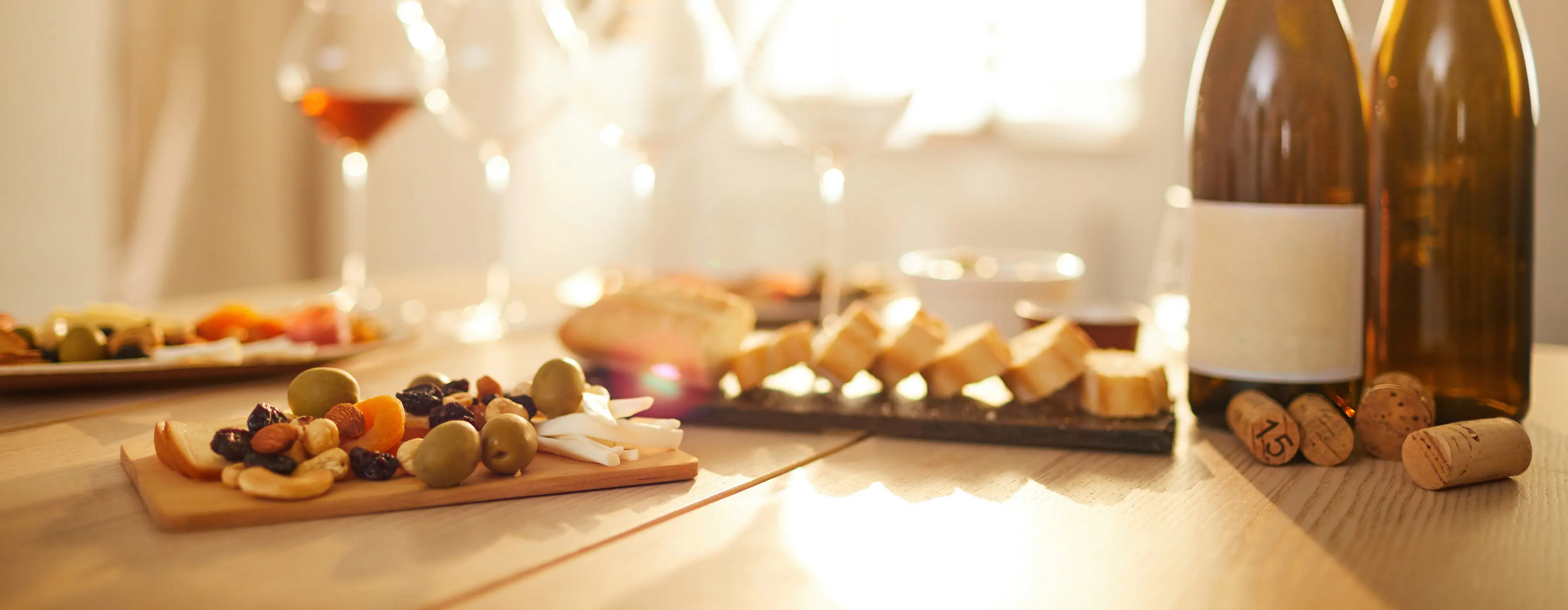 Foto em close de uma mesa com uma porção de castanhas, queijos e azeitonas. Ao fundo, outras porções, taças e garrafas de vinho.
