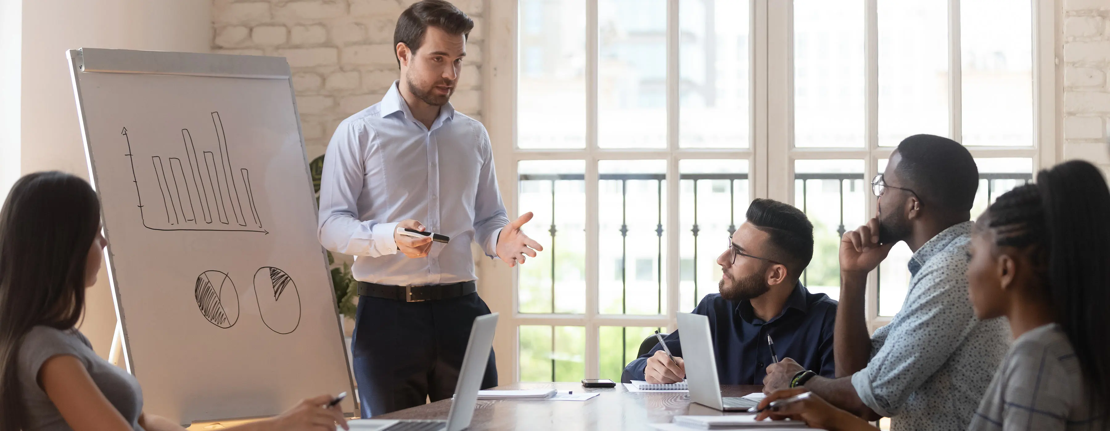 Foto de um homem jovem fazendo uma apresentação em pé de frente para um flip chart e algumas pessoas jovens sentadas ao redor de uma mesa prestando atenção.