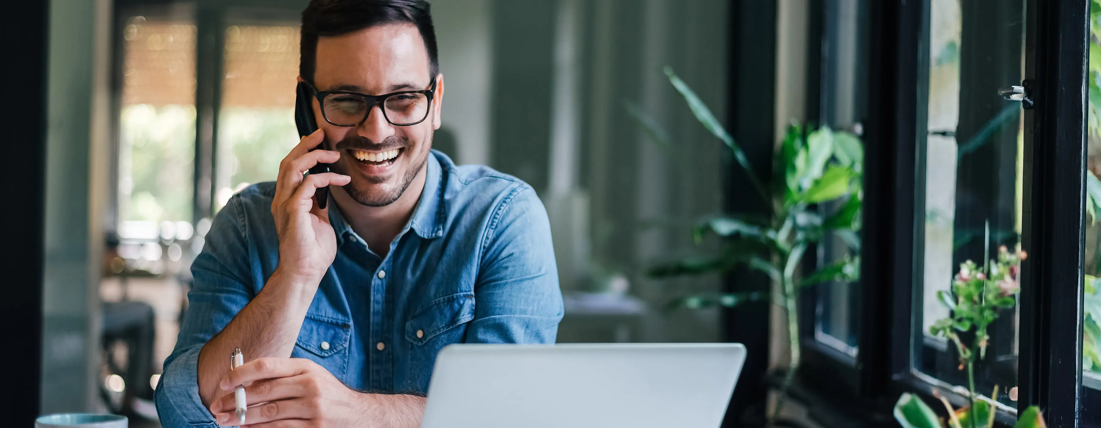 Foto de um homem jovem sorridente sentado em um escritório casual fazendo uma ligação com seu aparelho celular enquanto trabalha com o laptop em cima da mesa.