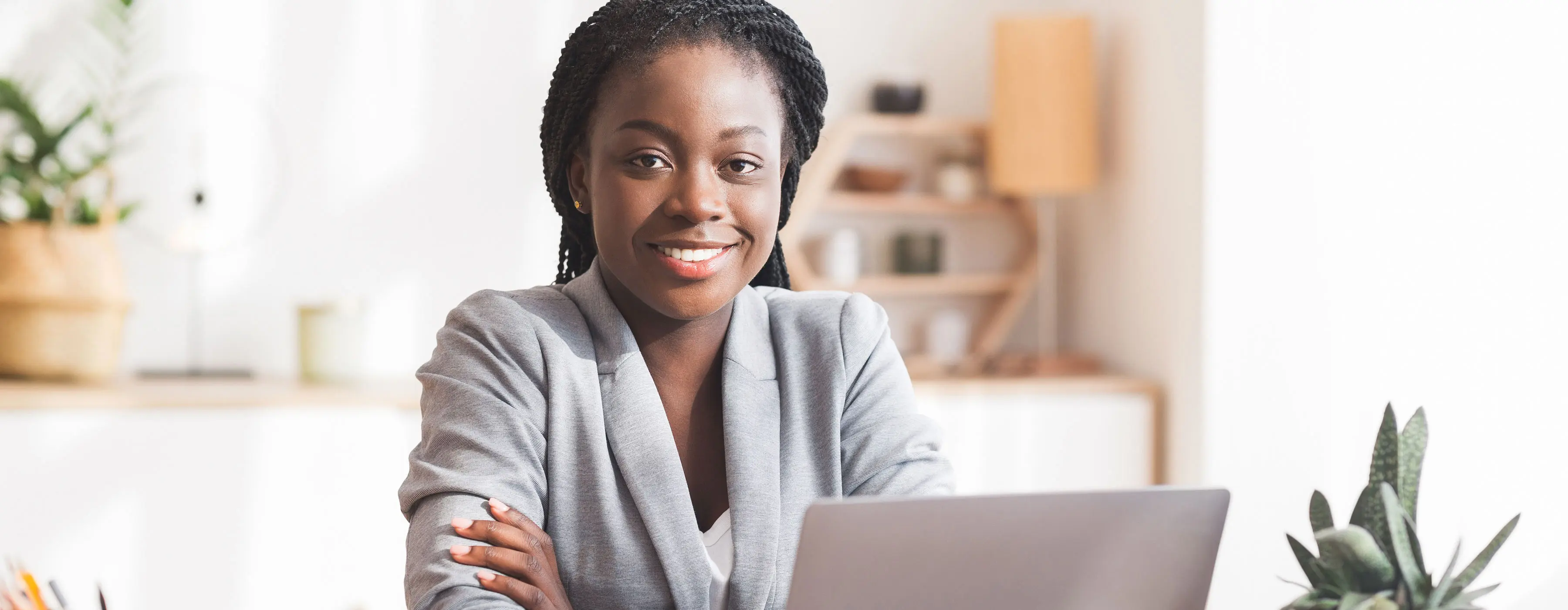 Foto de uma mulher jovem, sorridente e com roupas formais, fazendo pose para a foto, de frente para um notebook e sentada em um escritório.