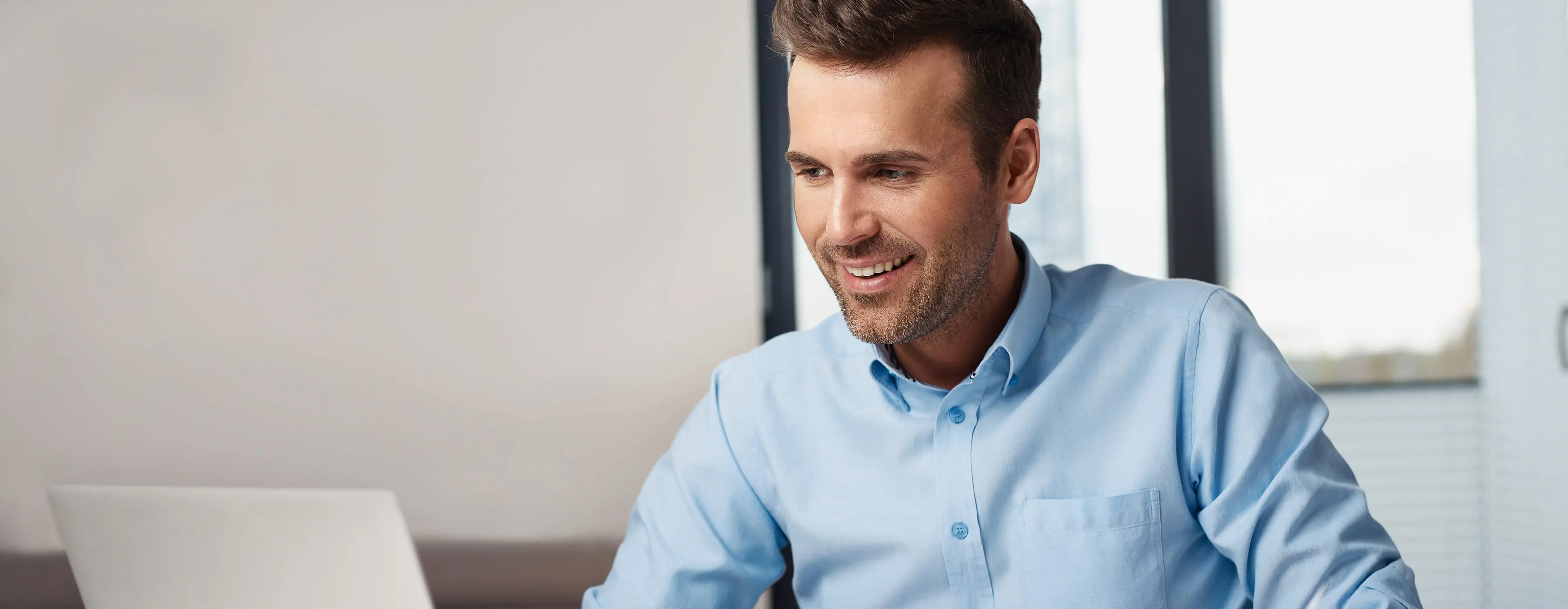 Foto de um homem jovem sorridente trabalhando home office e digitando em um notebook.