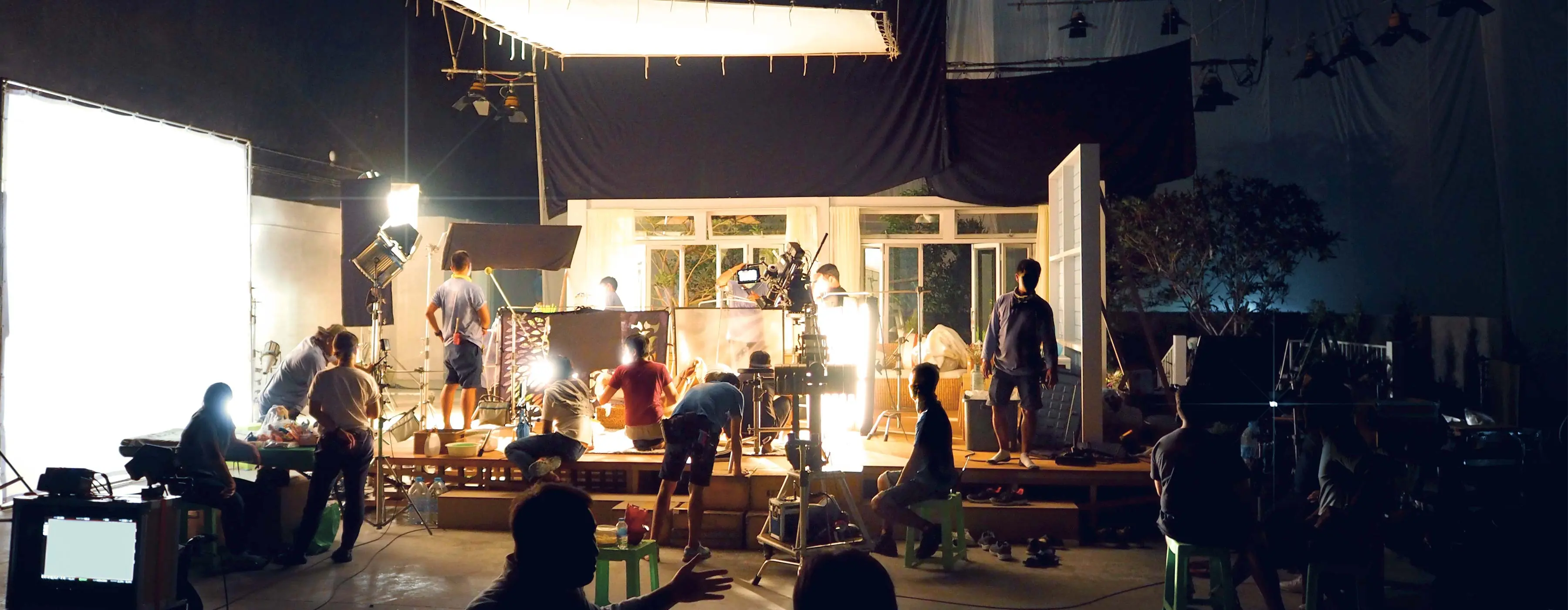 Foto de um grande estúdio de gravação e produção com um grupo de várias pessoas, cada uma em uma função diferente, mexendo em câmeras, organizando cabos, arrumando cenário e verificando iluminação.